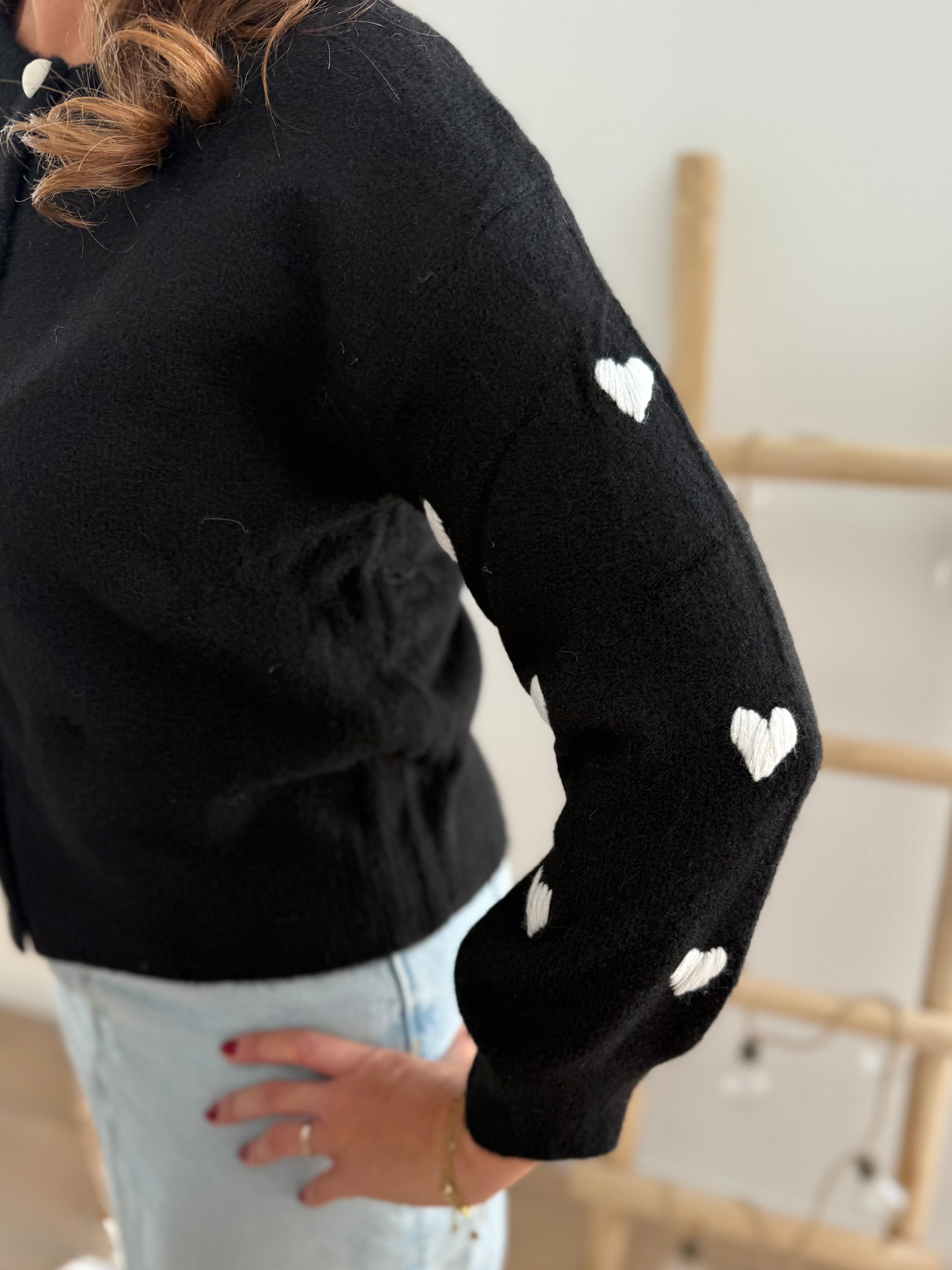 Strickjacke in schwarz mit weißen bestickten Herzen auf den Armen. Die Strickjacke hat einen Rundhalsausschnitt und eine Knopfleiste aus weißen Herzknöpfen.