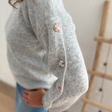 Grauer Strickpullover mit Rundhals. Knopfleiste an den armen aus pastelligem Stoff umzogenen Knöpfen.