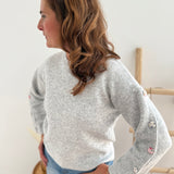 Grauer Strickpullover mit Rundhals. Knopfleiste an den armen aus pastelligem Stoff umzogenen Knöpfen.