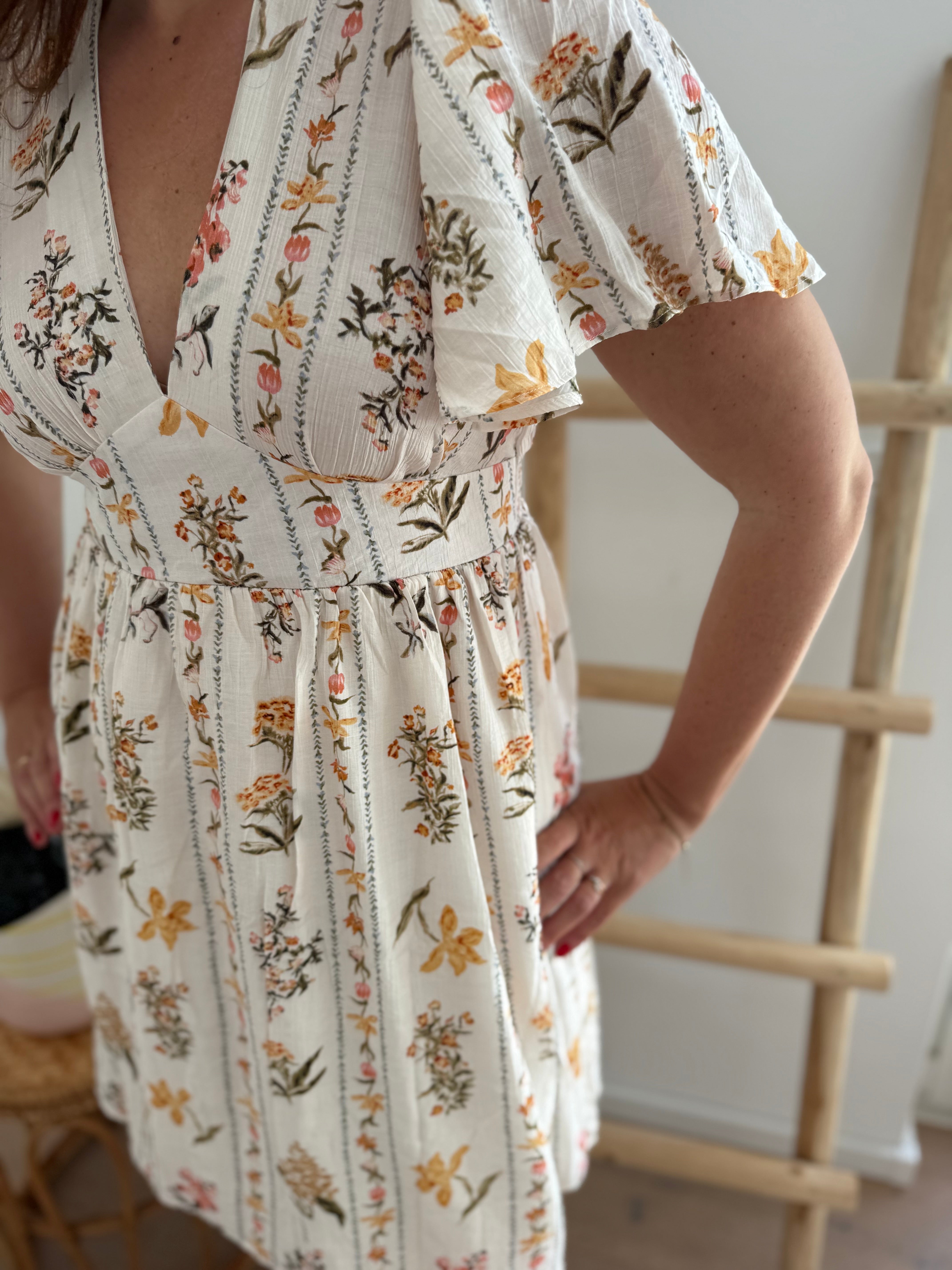 Leichtes kurzes Kleid mit V Ausschnitt und kurzen Flügelärmeln. Das Kleid ist cremefarben mit einem verspielten Blumenmuster in gelb, grün und rot.