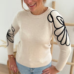 Beiger Strick Pullover mit Rundhalsausscnnitt und verspielter Blumenstickerei in schwarz an beiden Ärmeln.