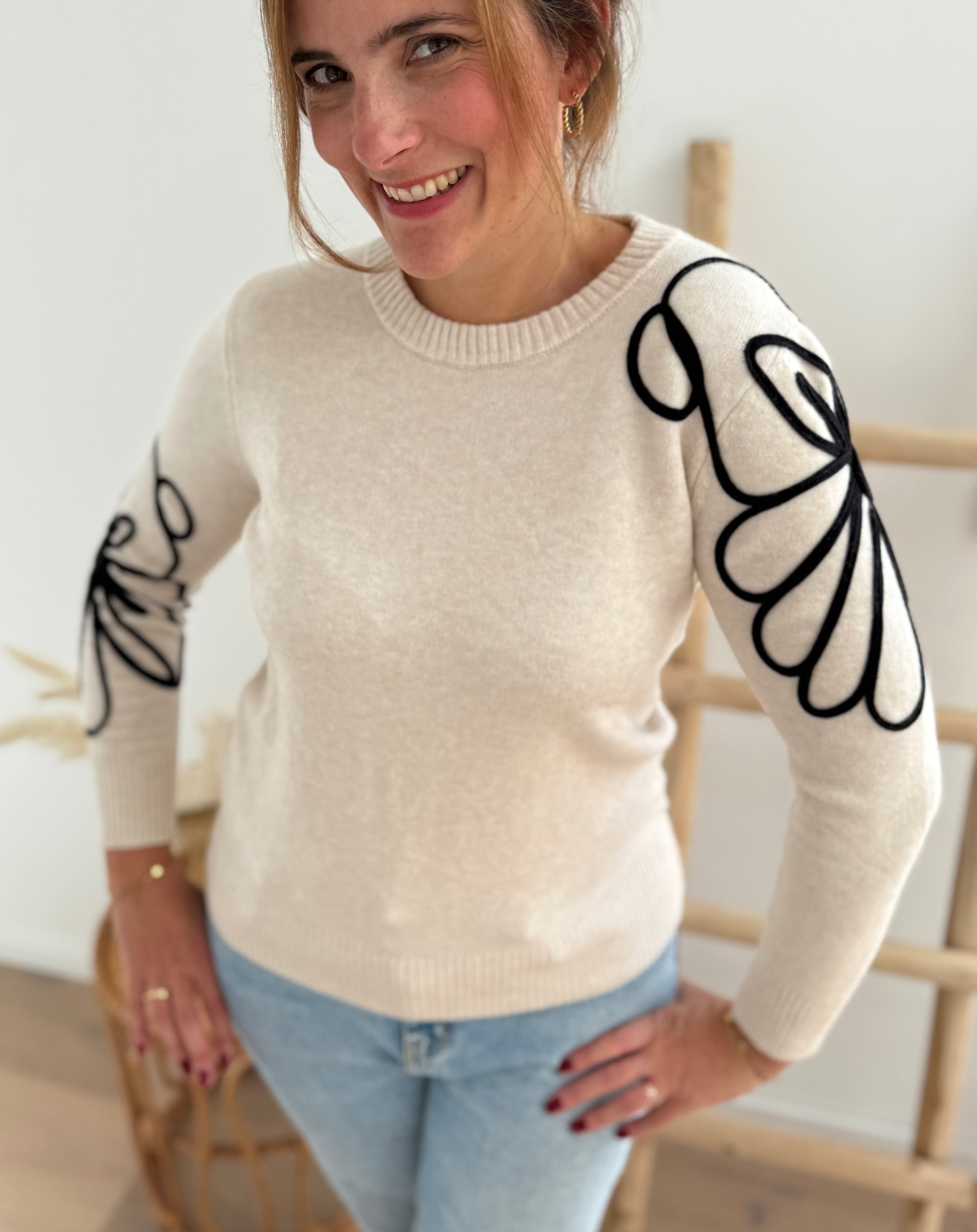 Beiger Strick Pullover mit Rundhalsausscnnitt und verspielter Blumenstickerei in schwarz an beiden Ärmeln.