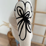 Beiger Strick Pullover mit Rundhalsausscnnitt und verspielter Blumenstickerei in schwarz an beiden Ärmeln.