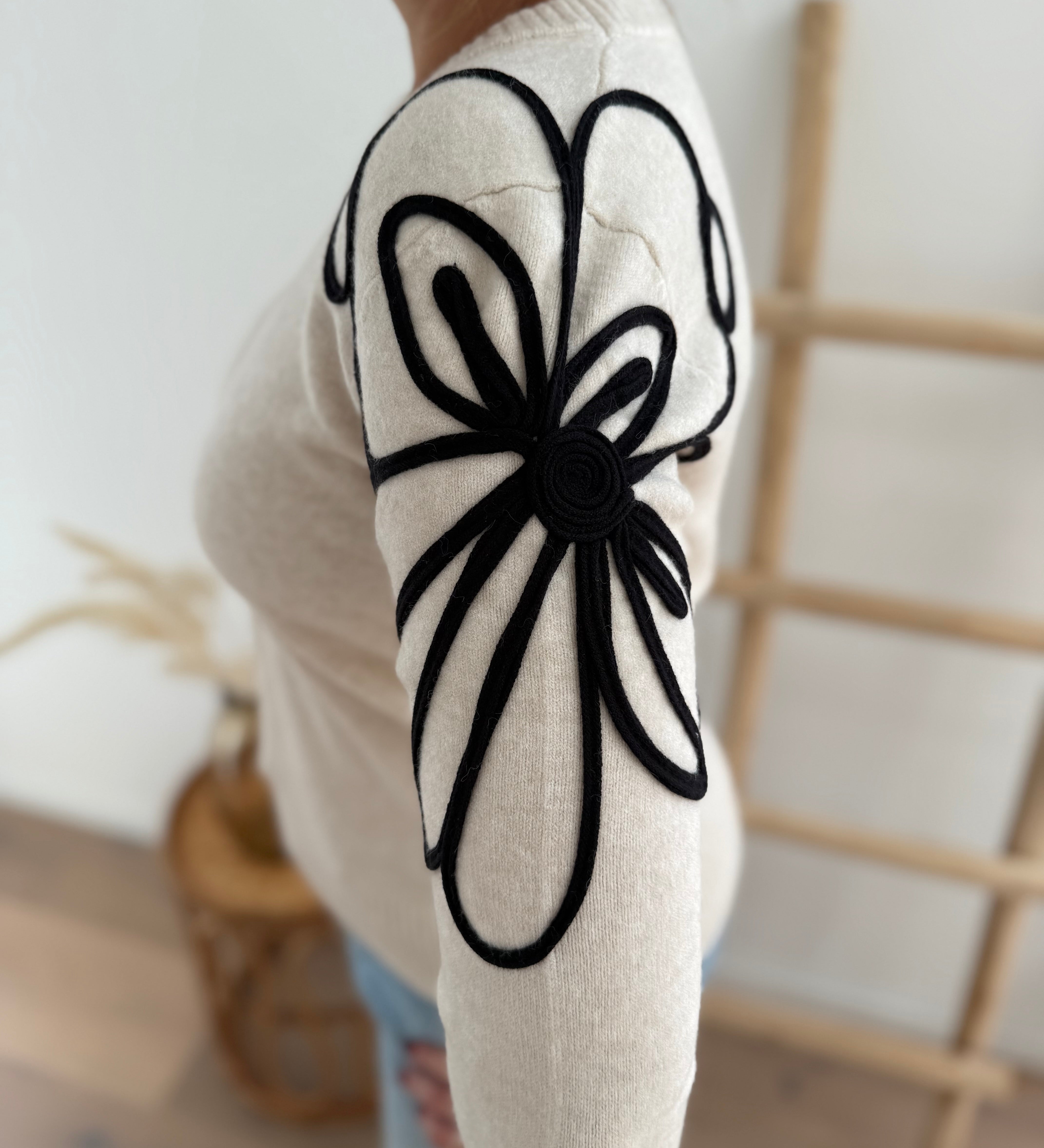 Beiger Strick Pullover mit Rundhalsausscnnitt und verspielter Blumenstickerei in schwarz an beiden Ärmeln.