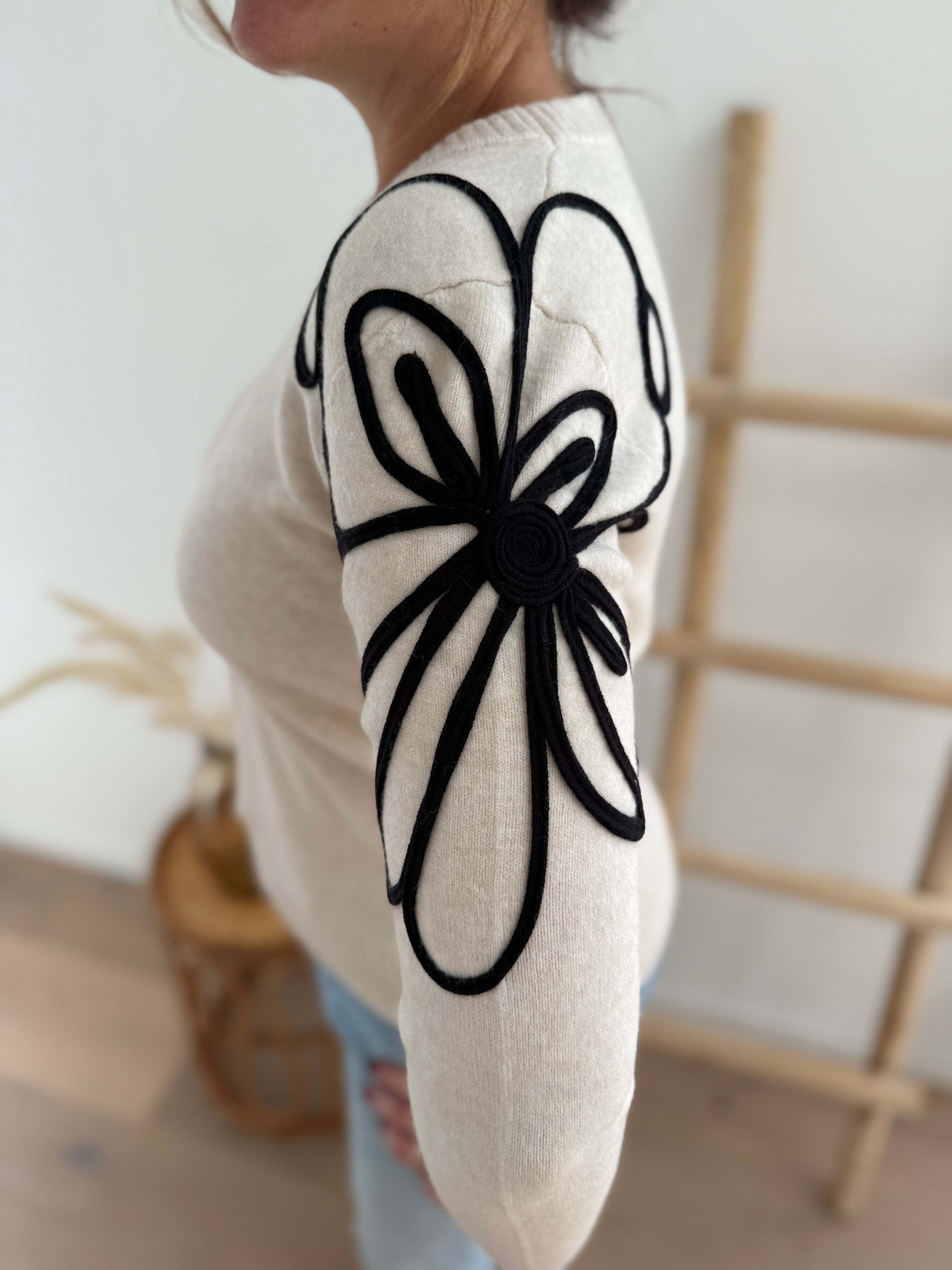 Beiger Strick Pullover mit Rundhalsausscnnitt und verspielter Blumenstickerei in schwarz an beiden Ärmeln.