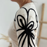 Beiger Strick Pullover mit Rundhalsausscnnitt und verspielter Blumenstickerei in schwarz an beiden Ärmeln.