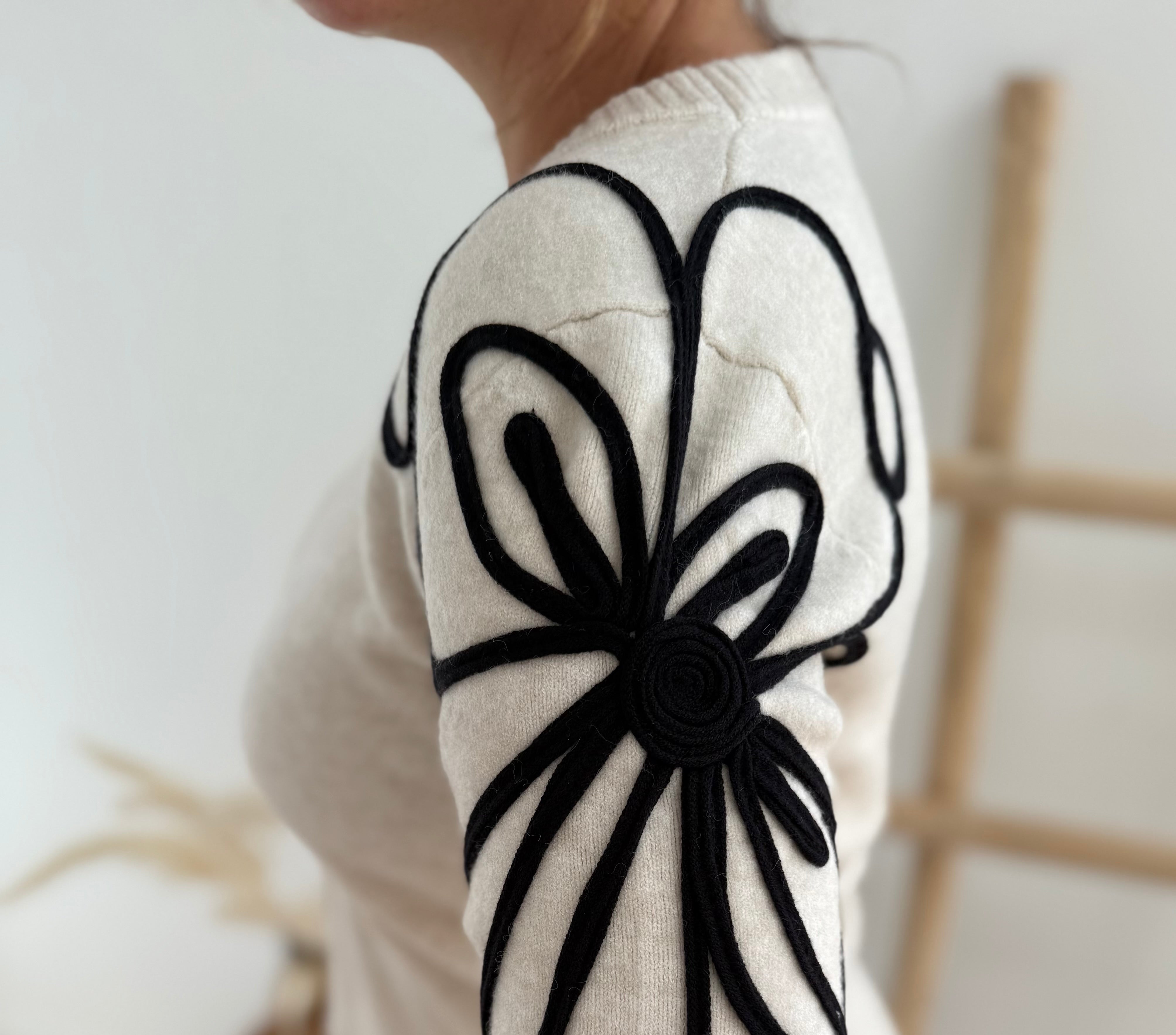 Beiger Strick Pullover mit Rundhalsausscnnitt und verspielter Blumenstickerei in schwarz an beiden Ärmeln.