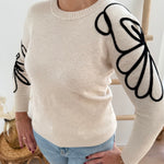 Beiger Strick Pullover mit Rundhalsausscnnitt und verspielter Blumenstickerei in schwarz an beiden Ärmeln.