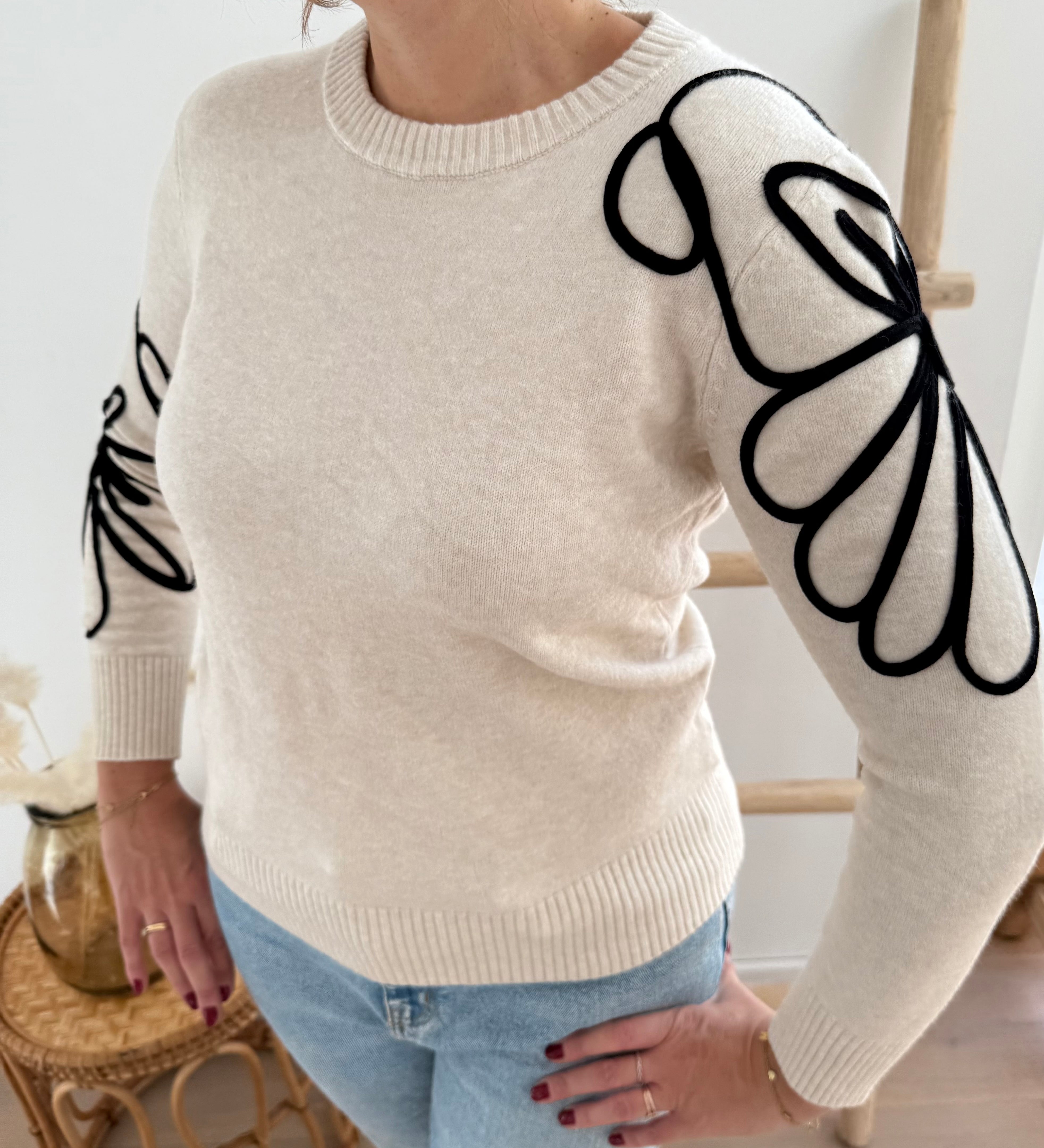Beiger Strick Pullover mit Rundhalsausscnnitt und verspielter Blumenstickerei in schwarz an beiden Ärmeln.