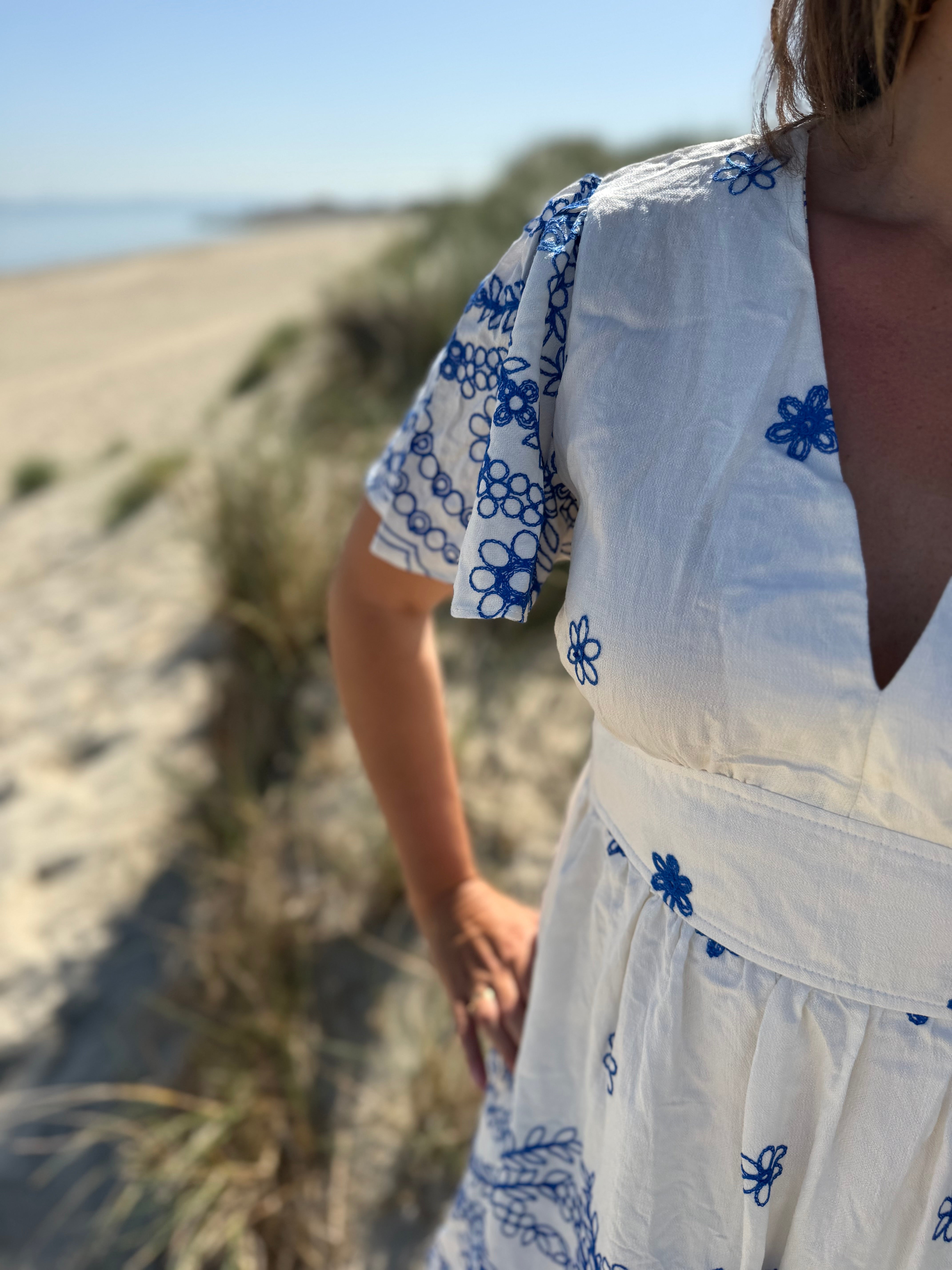 Leichtes kurzes Kleid mit V Ausschnitt und kurzen Flügelärmeln. Das Kleid ist weiß mit blauen Blumenstickereien.