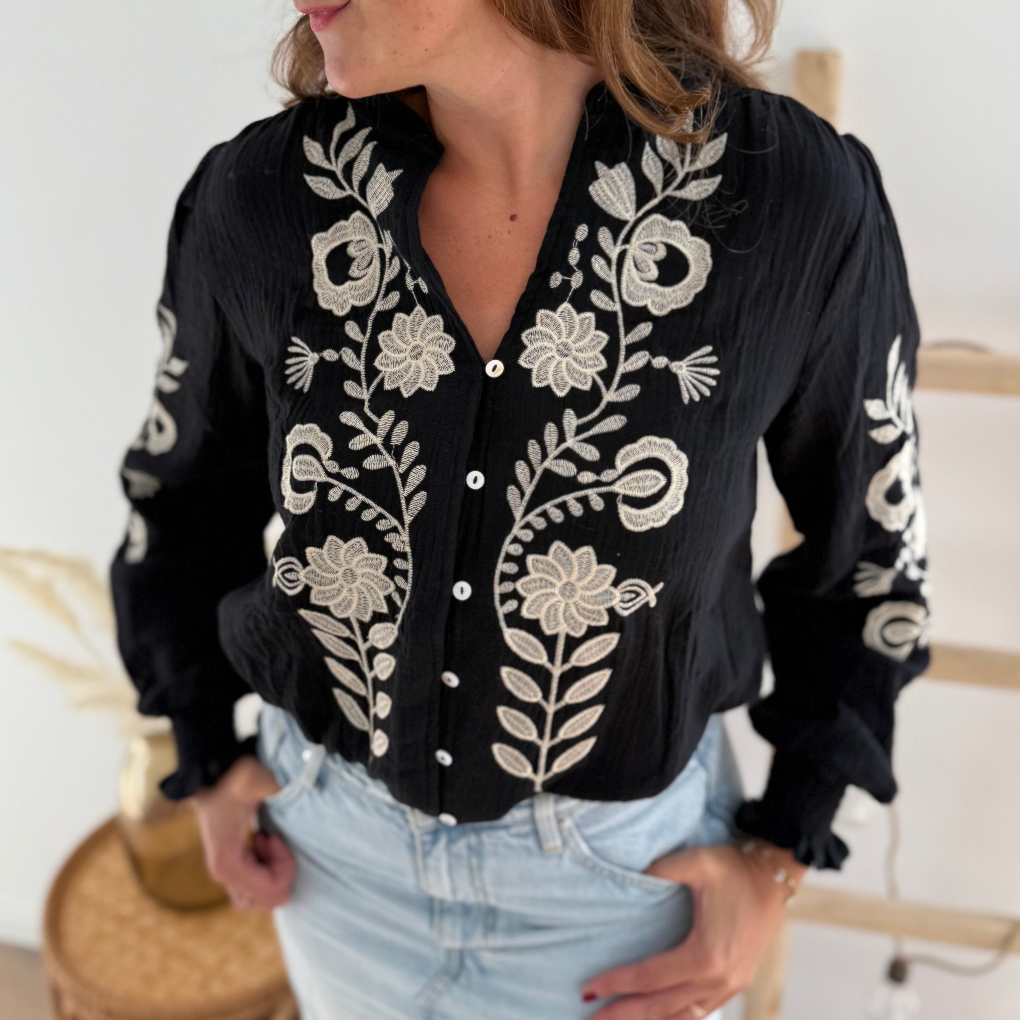 Schwarze Musselinbluse mit verspielten Blumenstickereien in beige vorne und auf den Armen. Die Bluse hat einen V Ausschnitt mit stehendem Rücschenkragen.