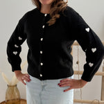 Strickjacke in schwarz mit weißen bestickten Herzen auf den Armen. Die Strickjacke hat einen Rundhalsausschnitt und eine Knopfleiste aus weißen Herzknöpfen.