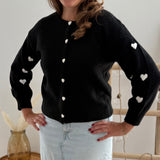 Strickjacke in schwarz mit weißen bestickten Herzen auf den Armen. Die Strickjacke hat einen Rundhalsausschnitt und eine Knopfleiste aus weißen Herzknöpfen.