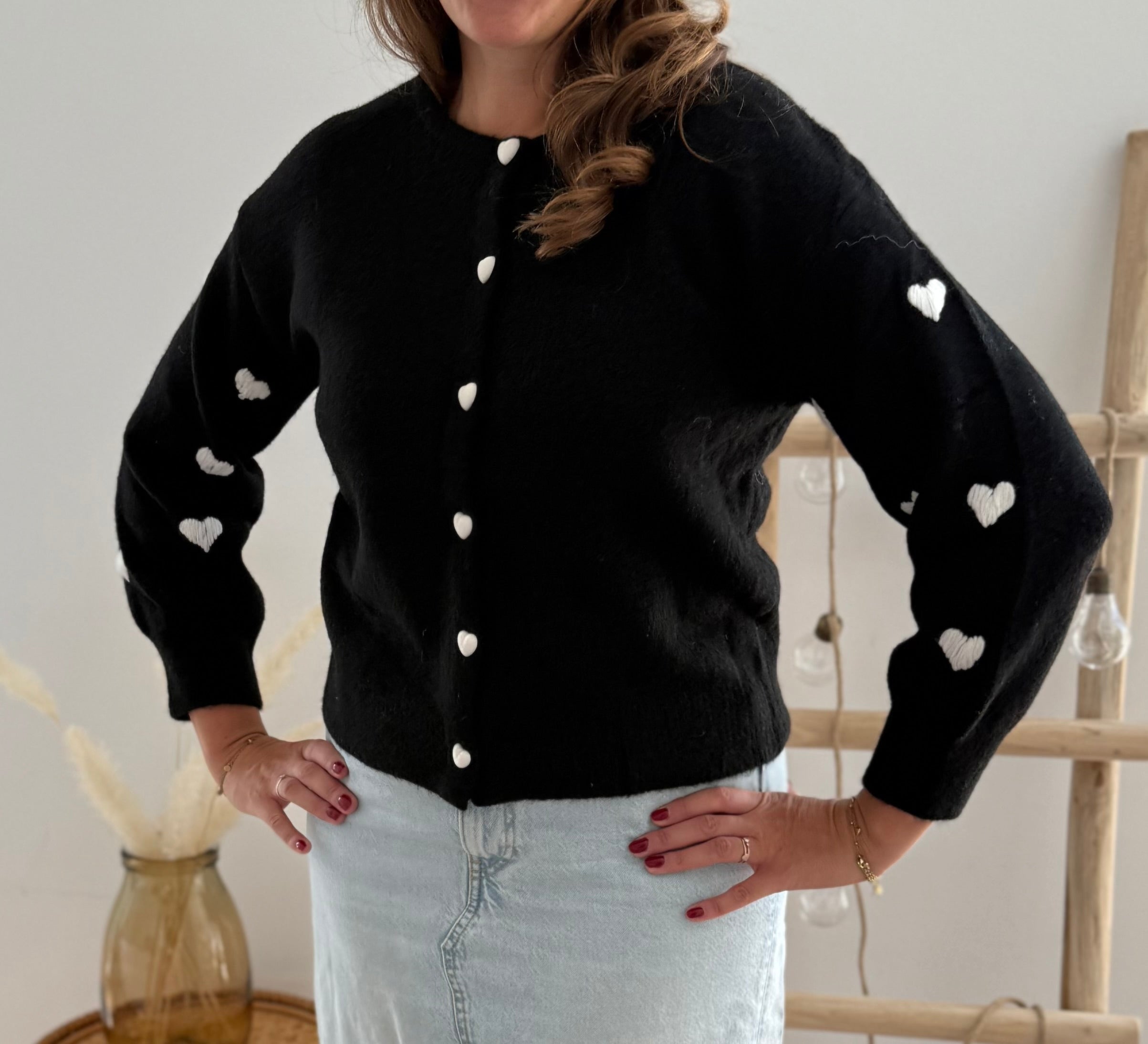 Strickjacke in schwarz mit weißen bestickten Herzen auf den Armen. Die Strickjacke hat einen Rundhalsausschnitt und eine Knopfleiste aus weißen Herzknöpfen.