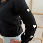 Strickjacke in schwarz mit weißen bestickten Herzen auf den Armen. Die Strickjacke hat einen Rundhalsausschnitt und eine Knopfleiste aus weißen Herzknöpfen.