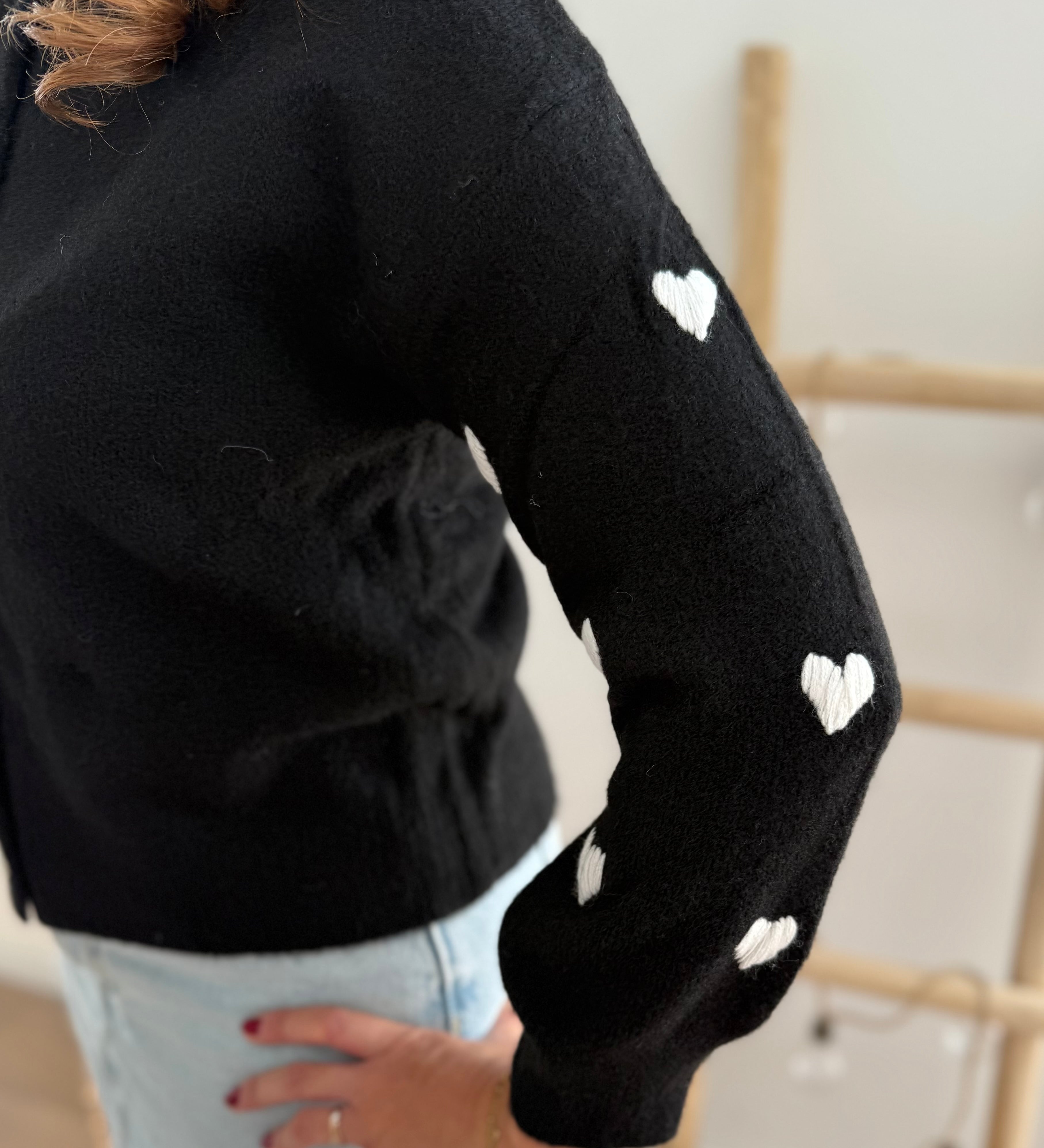 Strickjacke in schwarz mit weißen bestickten Herzen auf den Armen. Die Strickjacke hat einen Rundhalsausschnitt und eine Knopfleiste aus weißen Herzknöpfen.