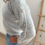 Grauer Strickpullover mit Rundhals. Knopfleiste an den armen aus pastelligem Stoff umzogenen Knöpfen.