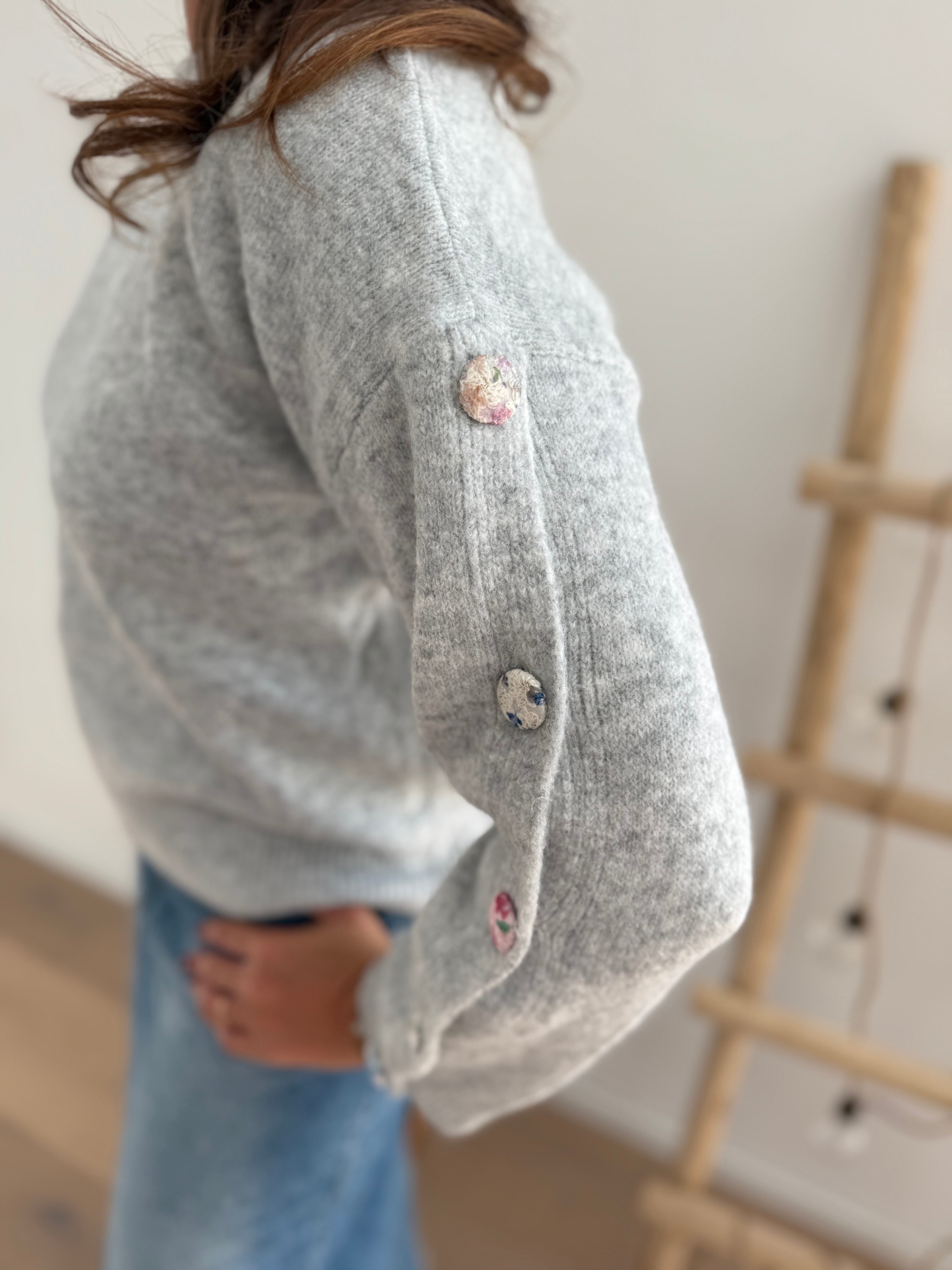 Grauer Strickpullover mit Rundhals. Knopfleiste an den armen aus pastelligem Stoff umzogenen Knöpfen.