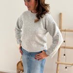Grauer Strickpullover mit Rundhals. Knopfleiste an den armen aus pastelligem Stoff umzogenen Knöpfen.