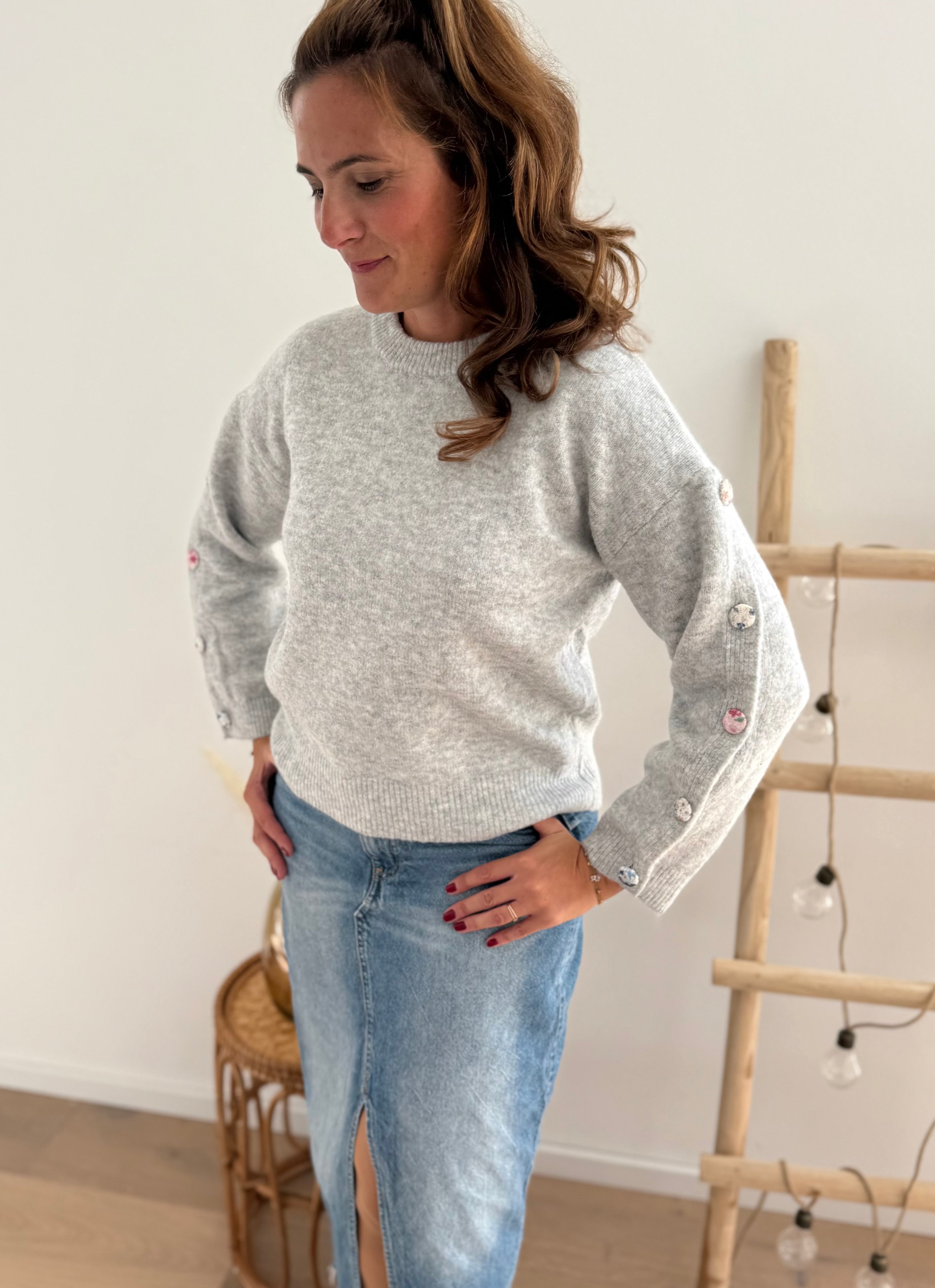 Grauer Strickpullover mit Rundhals. Knopfleiste an den armen aus pastelligem Stoff umzogenen Knöpfen.