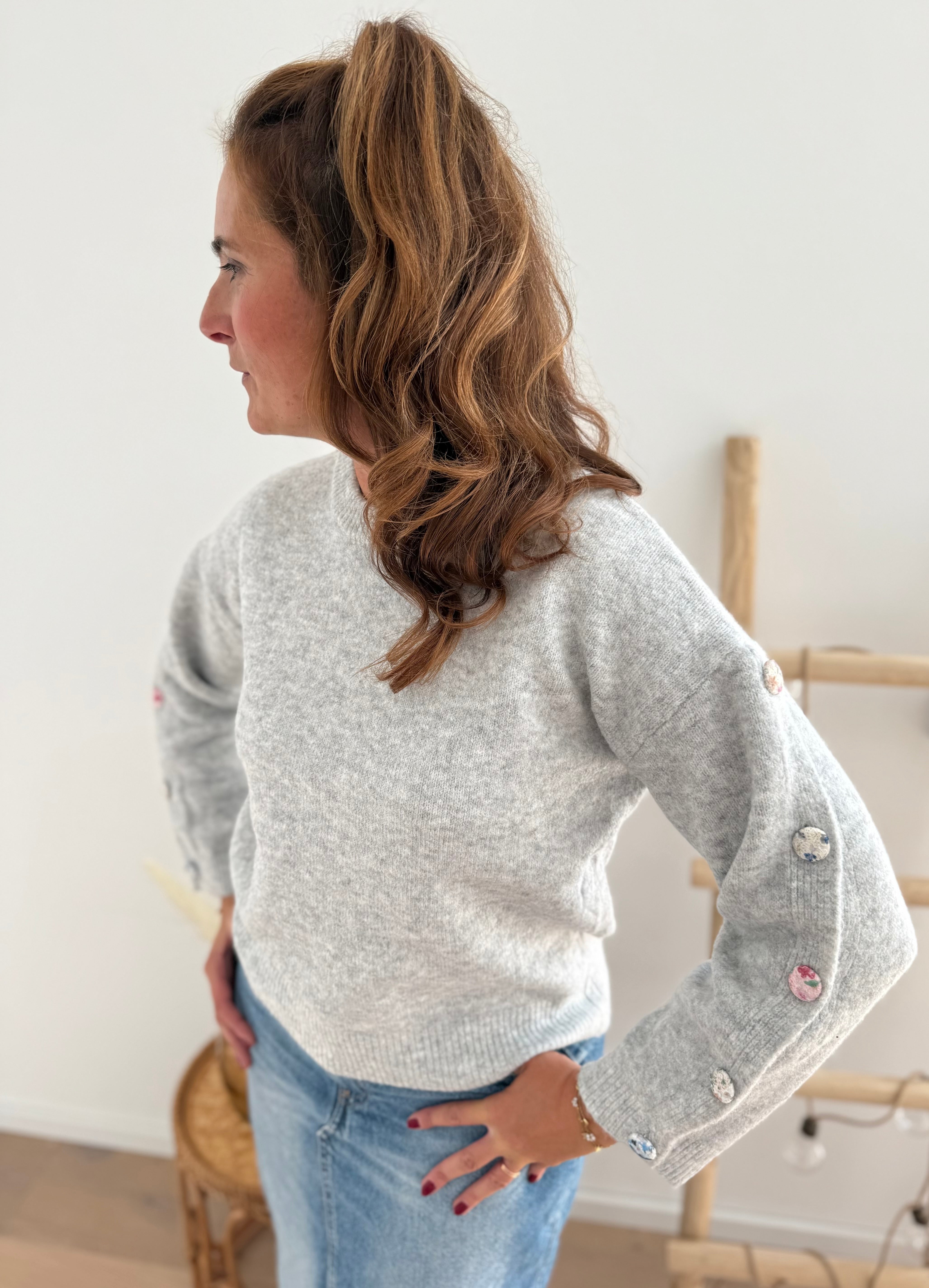 Grauer Strickpullover mit Rundhals. Knopfleiste an den armen aus pastelligem Stoff umzogenen Knöpfen.