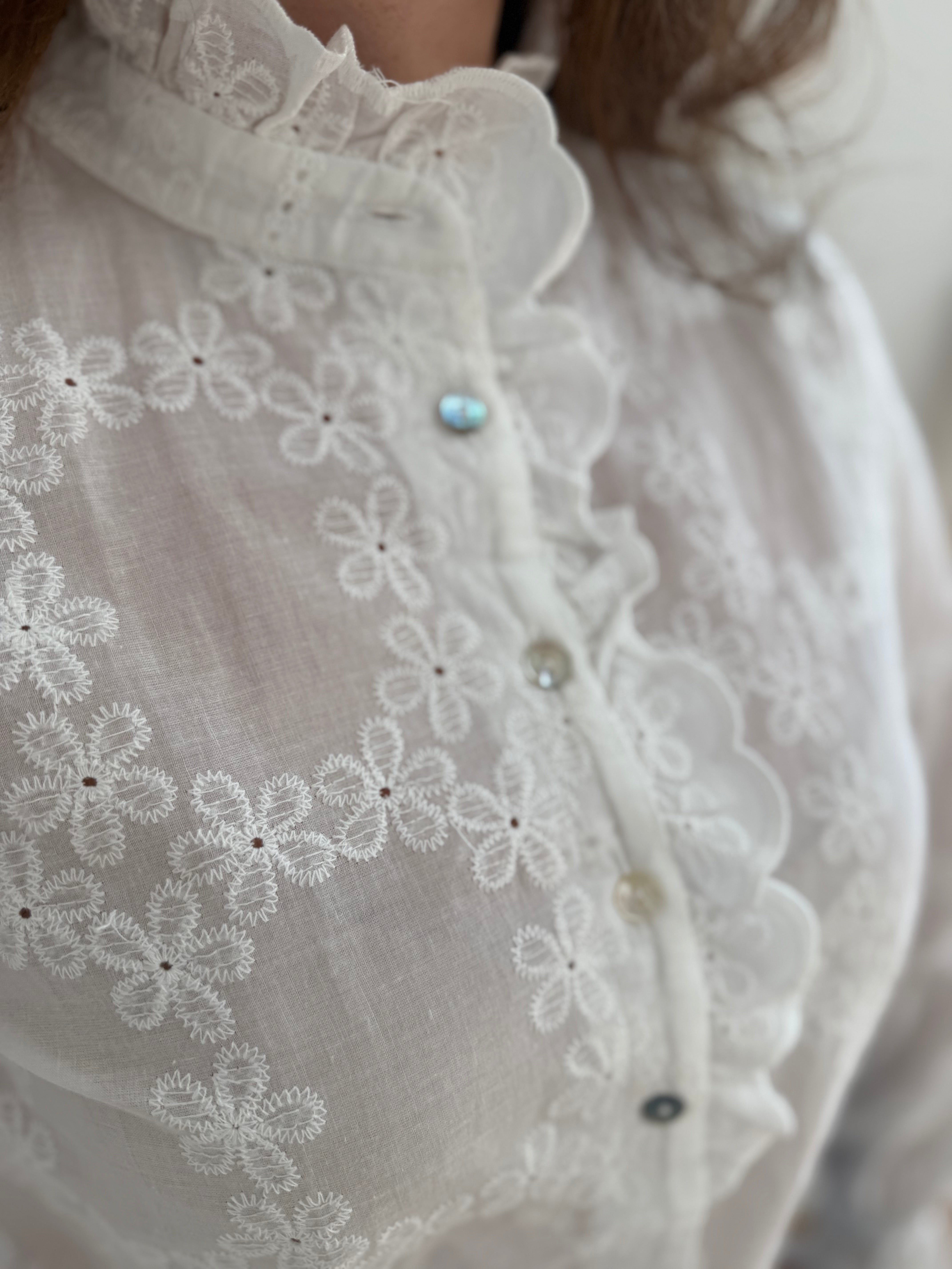 Weiße Bluse mit Blumen Stickereien. Die Bluse hat lange Ärmel, einen verspielter Stehkragen und eine Knopfleiste mit Spitze.