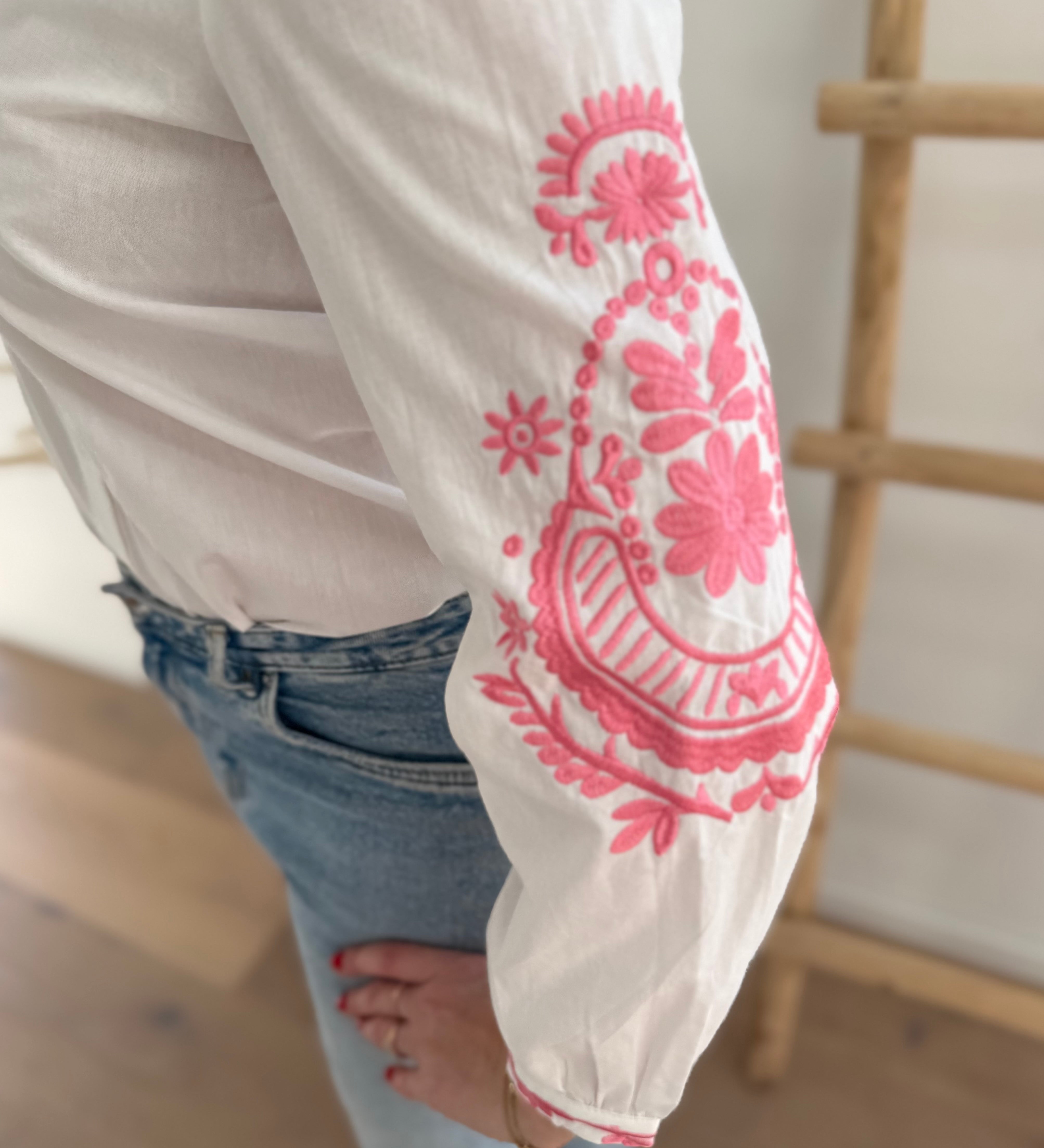 Weiße Bluse mit pinker Blumen Stickerei an den Ärmeln. Die Ärmel sind lang und der Kragen ist ein normaler Blusenkragen