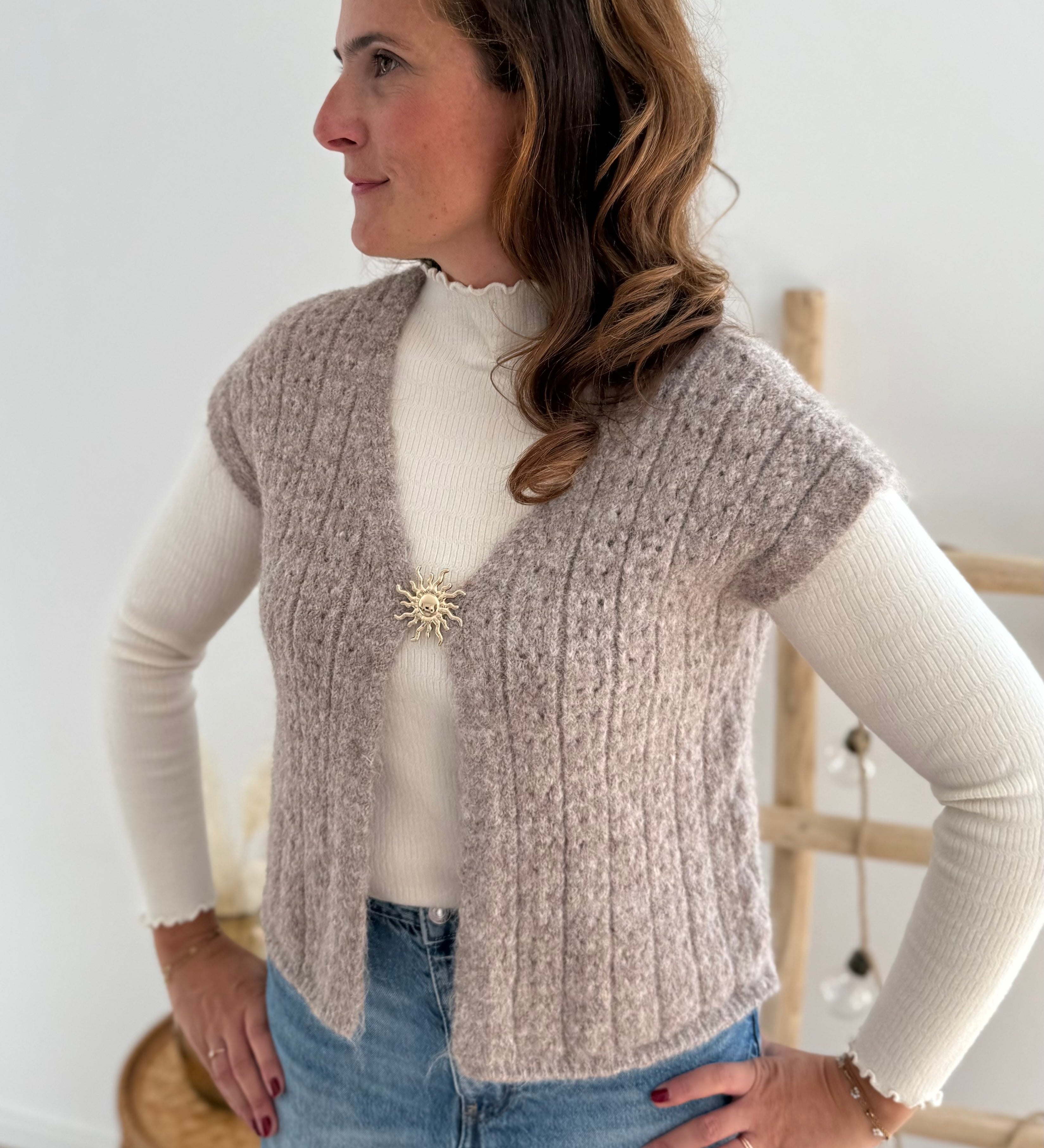 Locker fallende Strickweste in taupe. Die Weste wird von einer goldenen Brosche in Sonnenform zusammen gehalten.