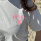 Oversize T-Shirt zum Verlieben. Kleines süßes Herz auf der Brust und ein großer Statement Print auf dem Rücken in rosa mit Amore. Das T-Shirt ist weiß.