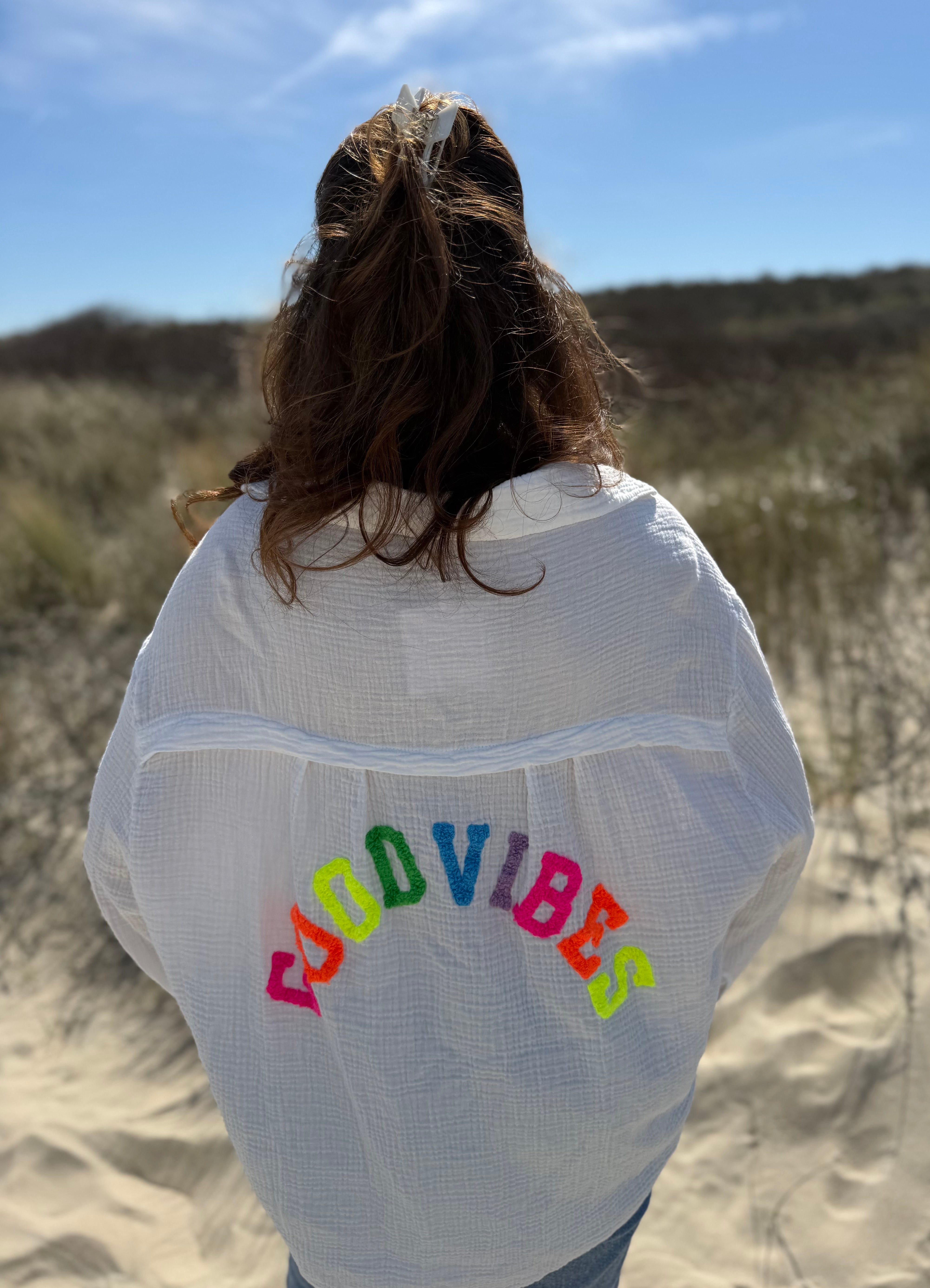 Musselinbluse im lockeren Oversize Look mit langen Ärmeln. Eyecatcher ist ein bunter GOOD VIBES Aufdruck am Rücken. Die Bluse ist weiß.