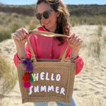Tasche aus naturfarbenem Bast mit weißem HELLO SUMMER Aufdruck. An dem Henkel hängt ein Anhänger aus bunten Quasten im Boho Look
