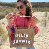 Tasche aus naturfarbenem Bast mit weißem HELLO SUMMER Aufdruck. An dem Henkel hängt ein Anhänger aus bunten Quasten im Boho Look