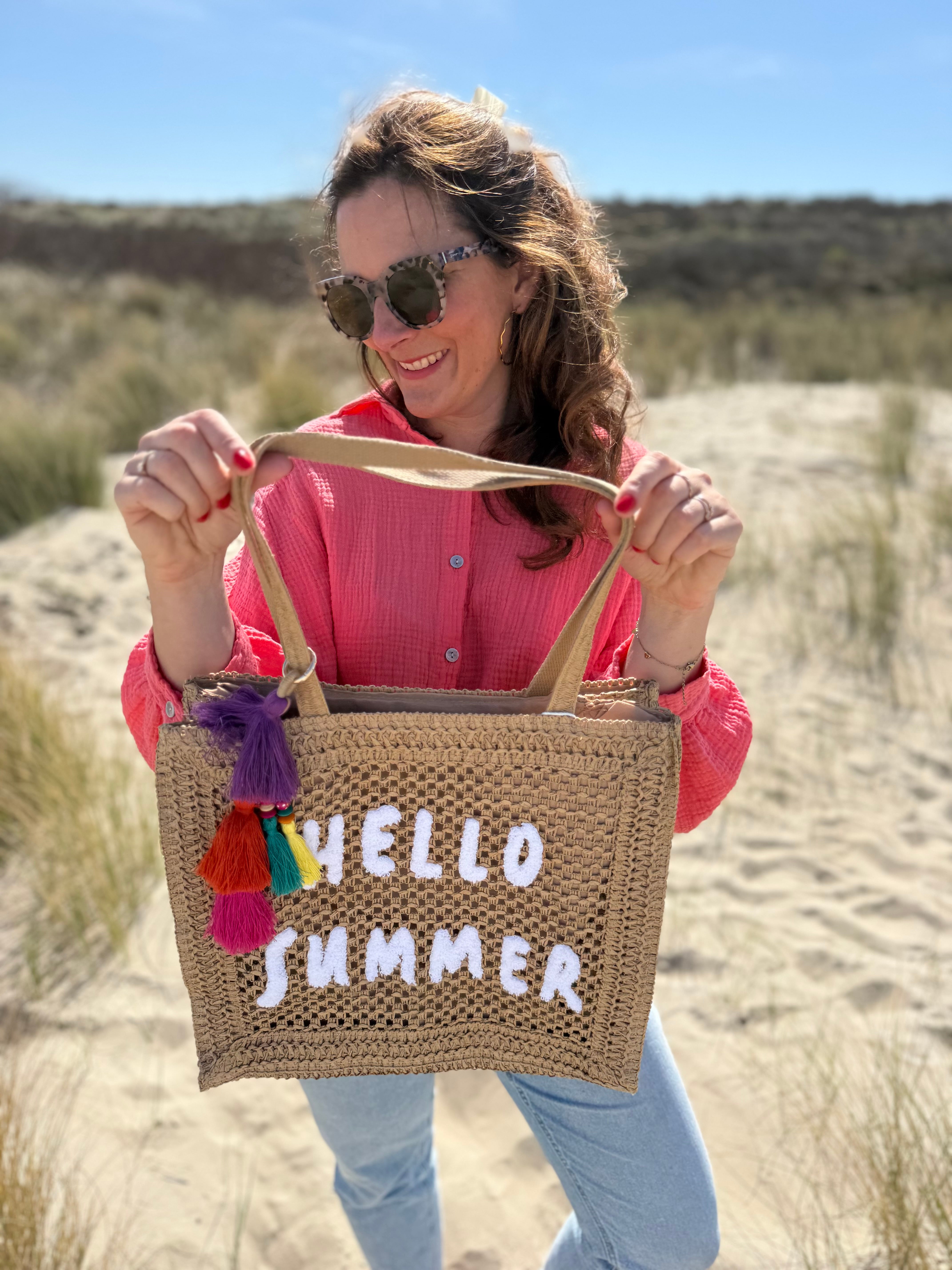 Tasche aus naturfarbenem Bast mit weißem HELLO SUMMER Aufdruck. An dem Henkel hängt ein Anhänger aus bunten Quasten im Boho Look