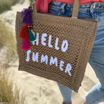 Tasche aus naturfarbenem Bast mit weißem HELLO SUMMER Aufdruck. An dem Henkel hängt ein Anhänger aus bunten Quasten im Boho Look