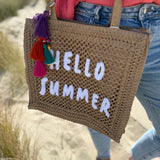Tasche aus naturfarbenem Bast mit weißem HELLO SUMMER Aufdruck. An dem Henkel hängt ein Anhänger aus bunten Quasten im Boho Look