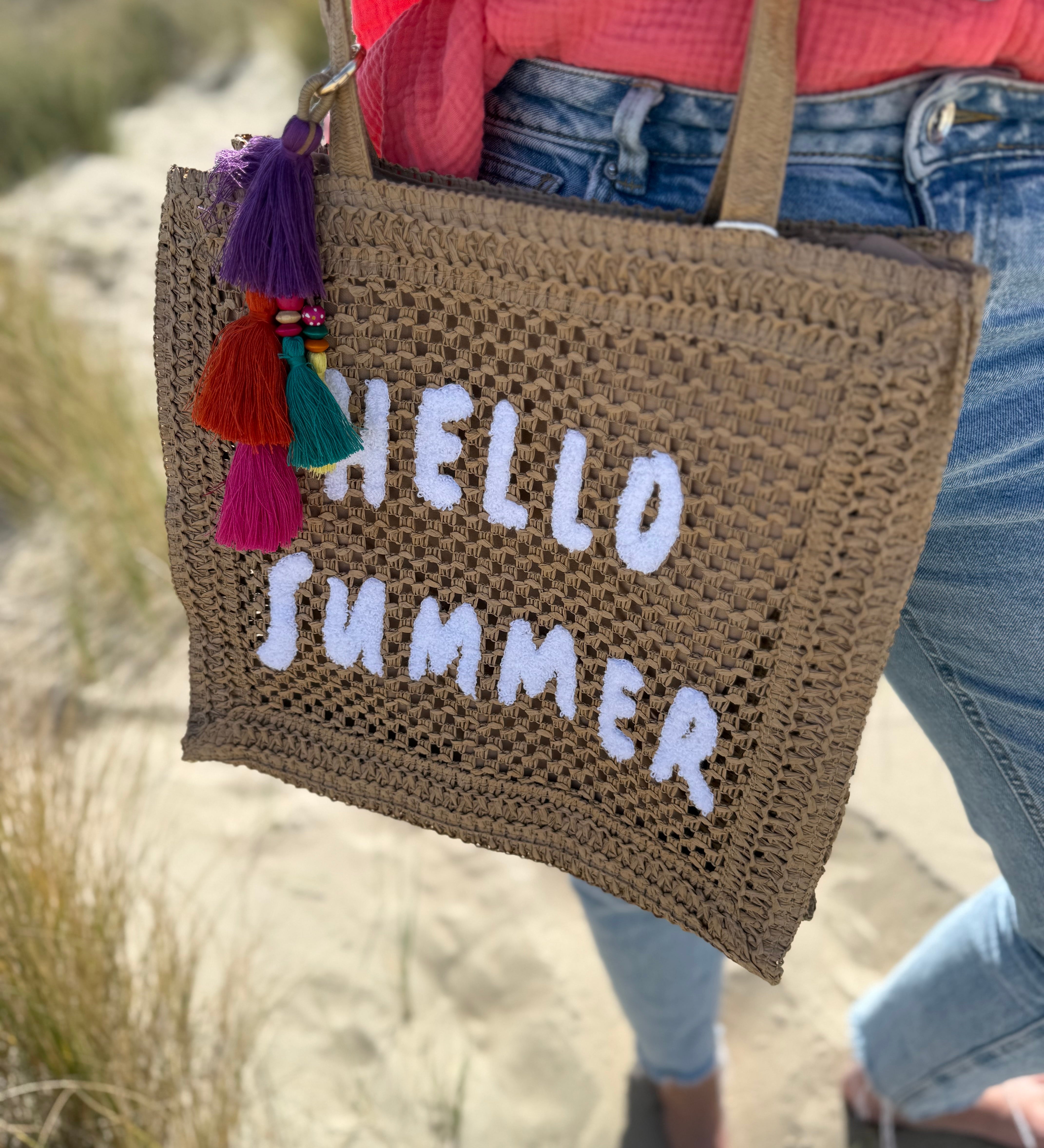 Tasche aus naturfarbenem Bast mit weißem HELLO SUMMER Aufdruck. An dem Henkel hängt ein Anhänger aus bunten Quasten im Boho Look