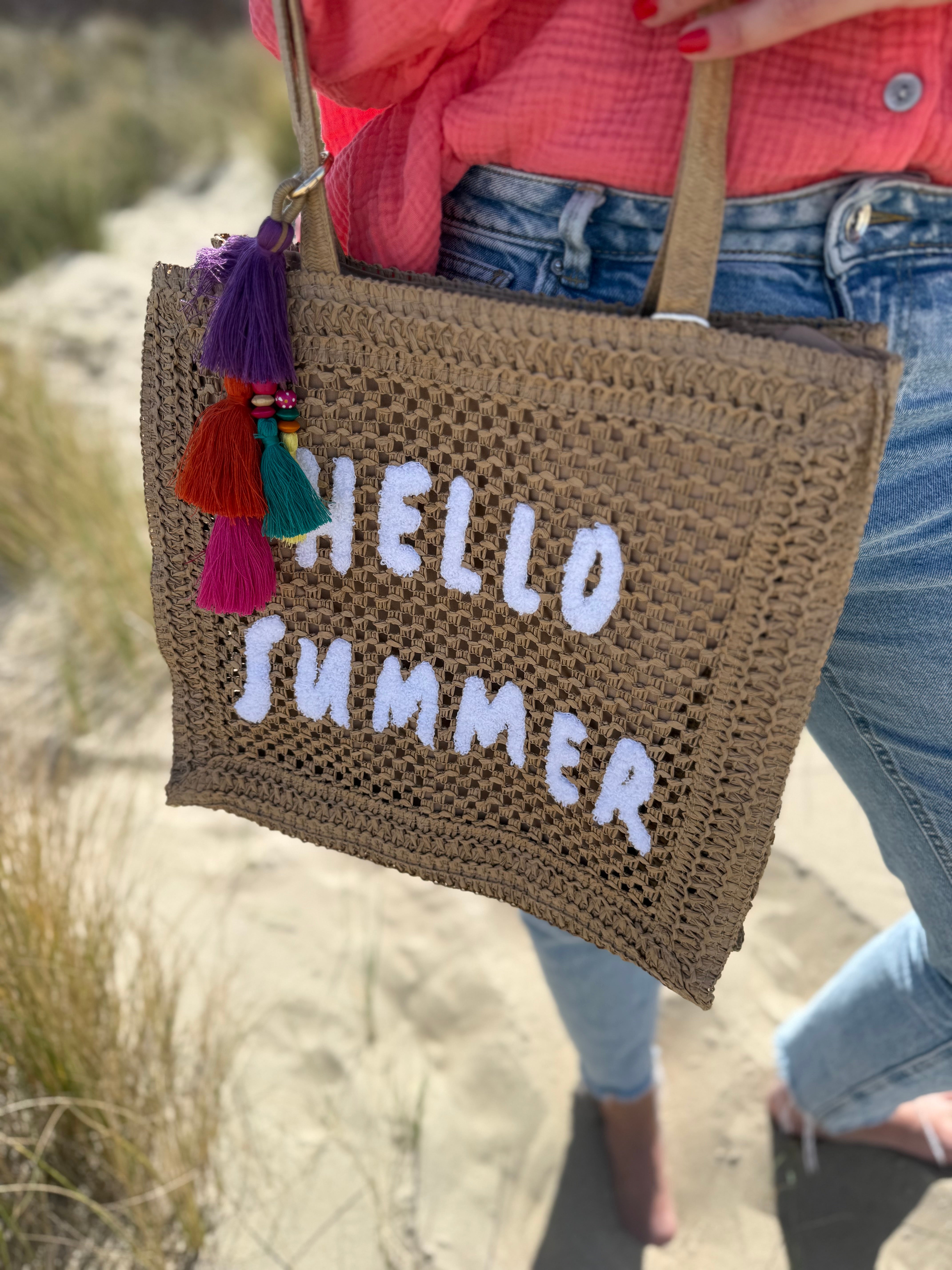 Tasche aus naturfarbenem Bast mit weißem HELLO SUMMER Aufdruck. An dem Henkel hängt ein Anhänger aus bunten Quasten im Boho Look
