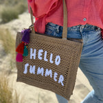Tasche aus naturfarbenem Bast mit weißem HELLO SUMMER Aufdruck. An dem Henkel hängt ein Anhänger aus bunten Quasten im Boho Look