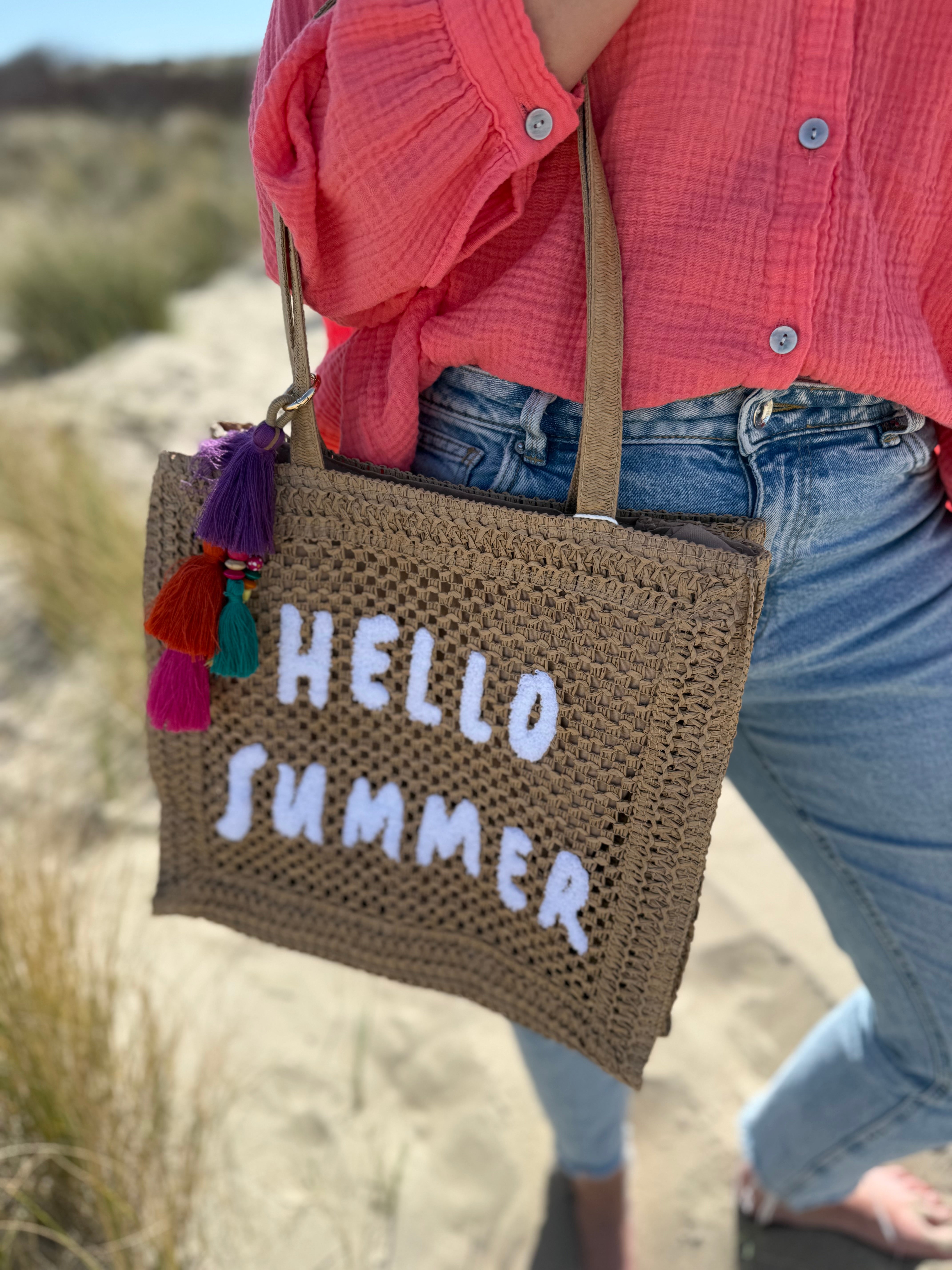 Tasche aus naturfarbenem Bast mit weißem HELLO SUMMER Aufdruck. An dem Henkel hängt ein Anhänger aus bunten Quasten im Boho Look