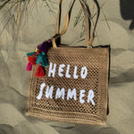 Tasche aus naturfarbenem Bast mit weißem HELLO SUMMER Aufdruck. An dem Henkel hängt ein Anhänger aus bunten Quasten im Boho Look