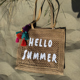 Tasche aus naturfarbenem Bast mit weißem HELLO SUMMER Aufdruck. An dem Henkel hängt ein Anhänger aus bunten Quasten im Boho Look
