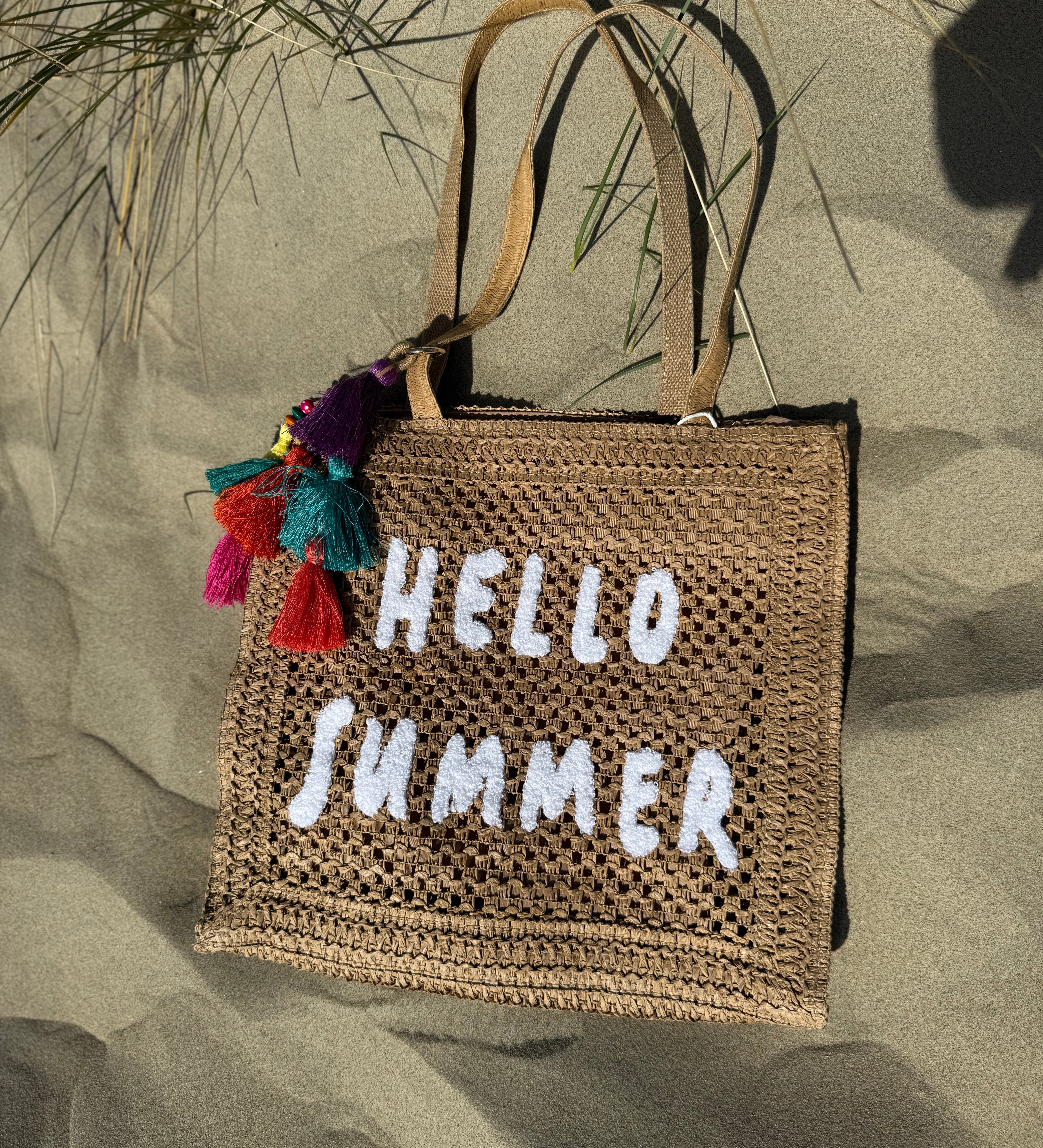 Tasche aus naturfarbenem Bast mit weißem HELLO SUMMER Aufdruck. An dem Henkel hängt ein Anhänger aus bunten Quasten im Boho Look