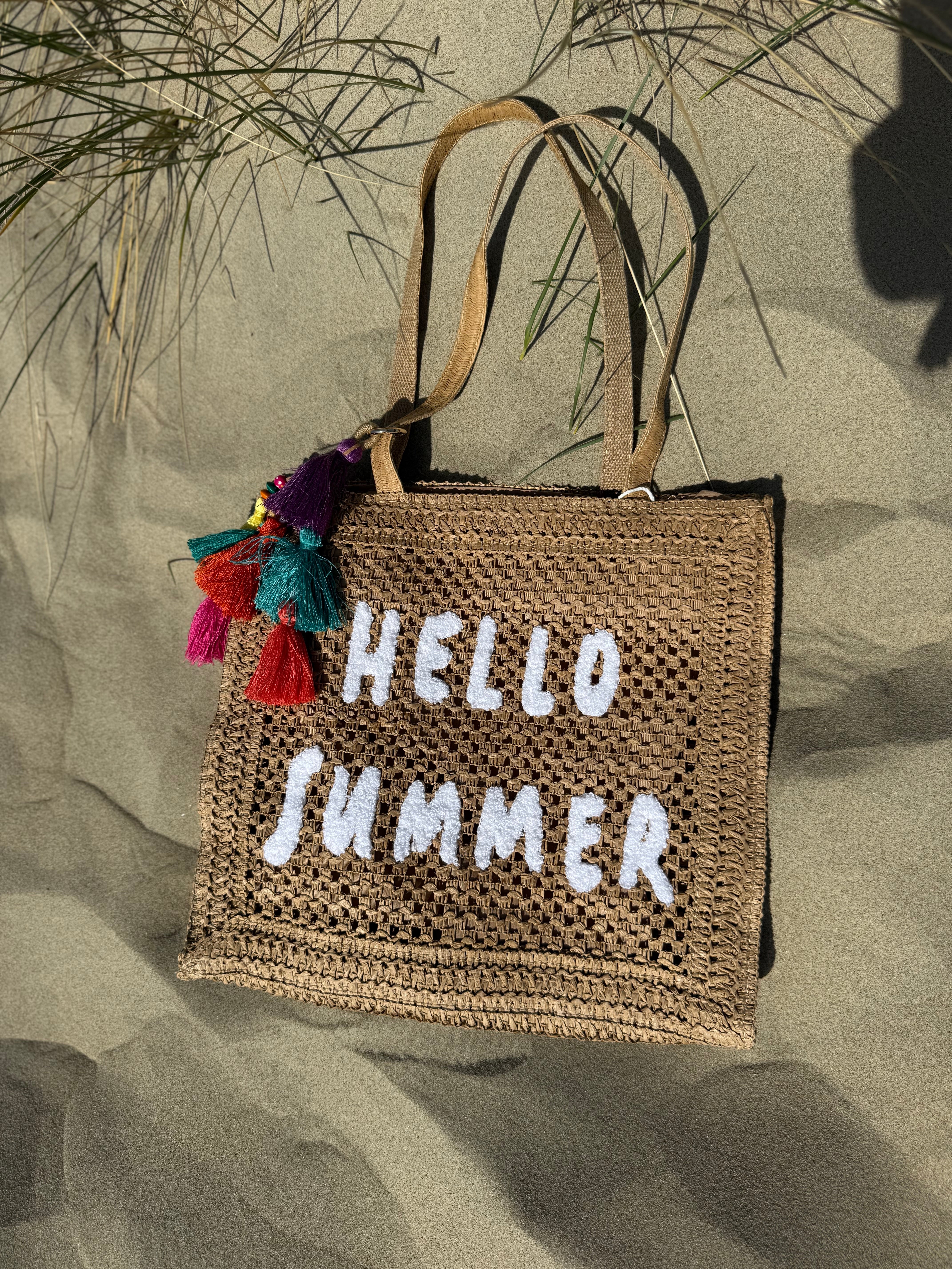 Tasche aus naturfarbenem Bast mit weißem HELLO SUMMER Aufdruck. An dem Henkel hängt ein Anhänger aus bunten Quasten im Boho Look