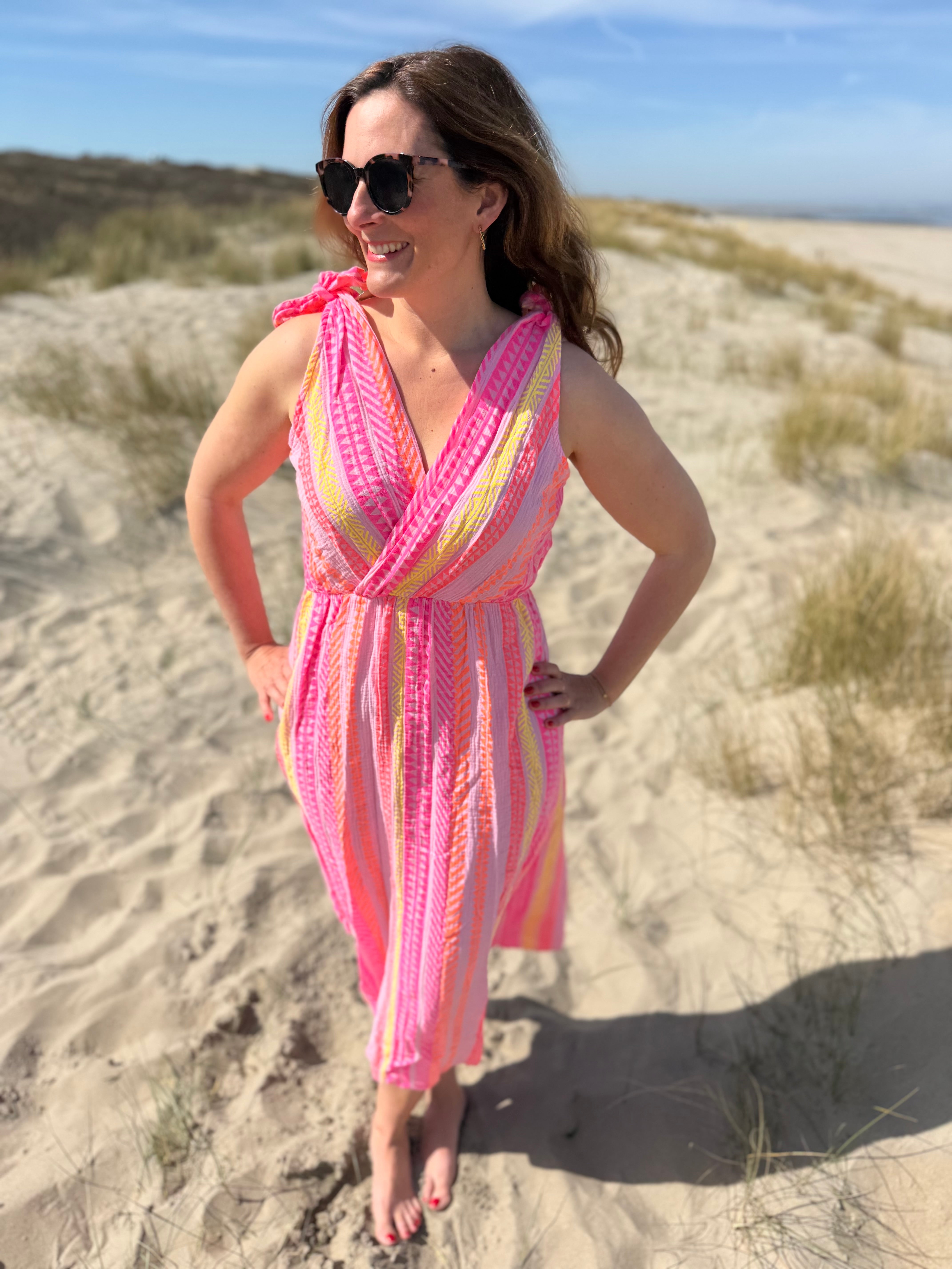 Musselinkleid in Midilänge mit V Ausschnitt. Es ist ärmellos und hat Schleifen an den Schultern zum binden. Das Kleid ist rosa mit pink, gelb und orangenen Ethno Muster.