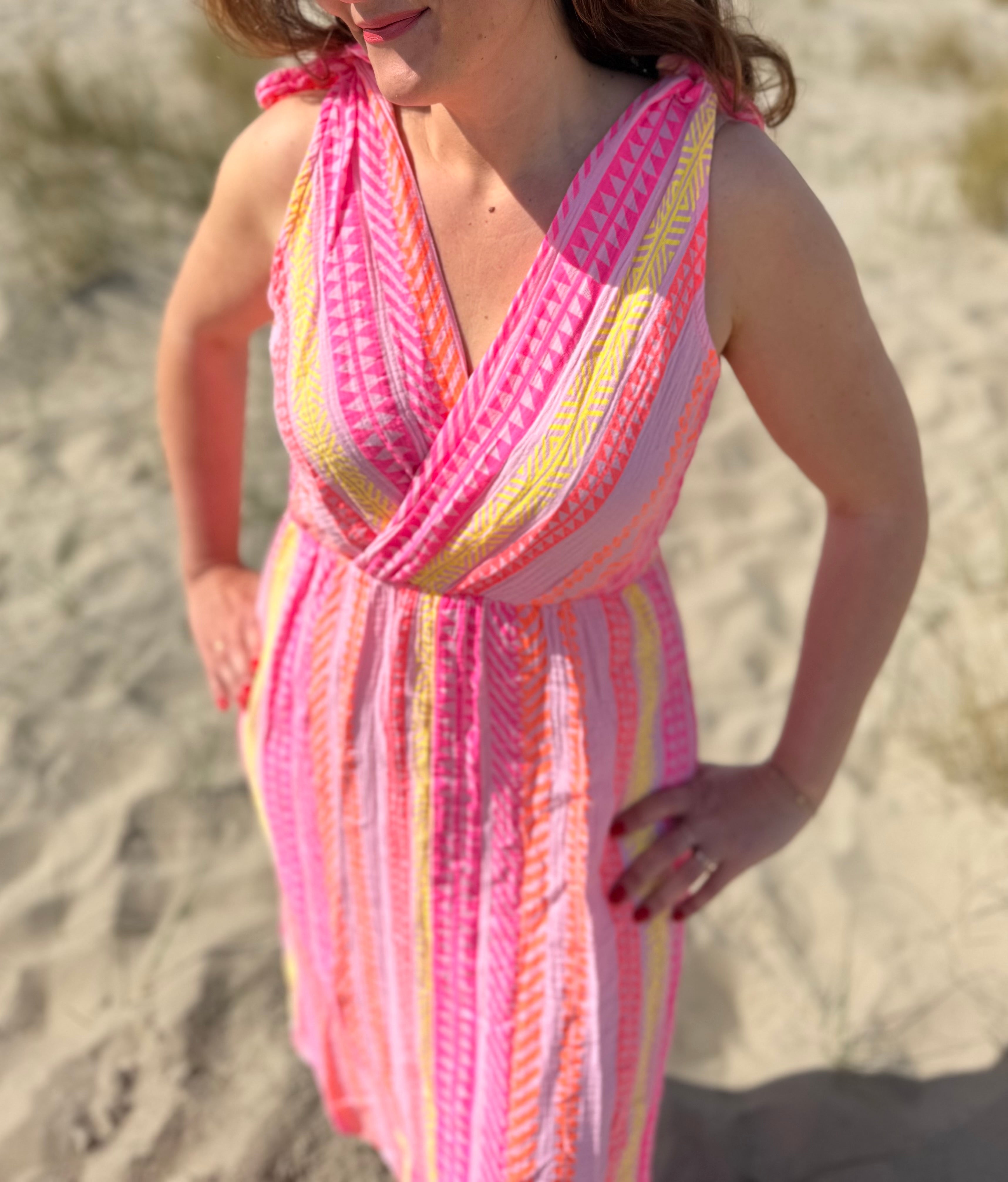 Musselinkleid in Midilänge mit V Ausschnitt. Es ist ärmellos und hat Schleifen an den Schultern zum binden. Das Kleid ist rosa mit pink, gelb und orangenen Ethno Muster.