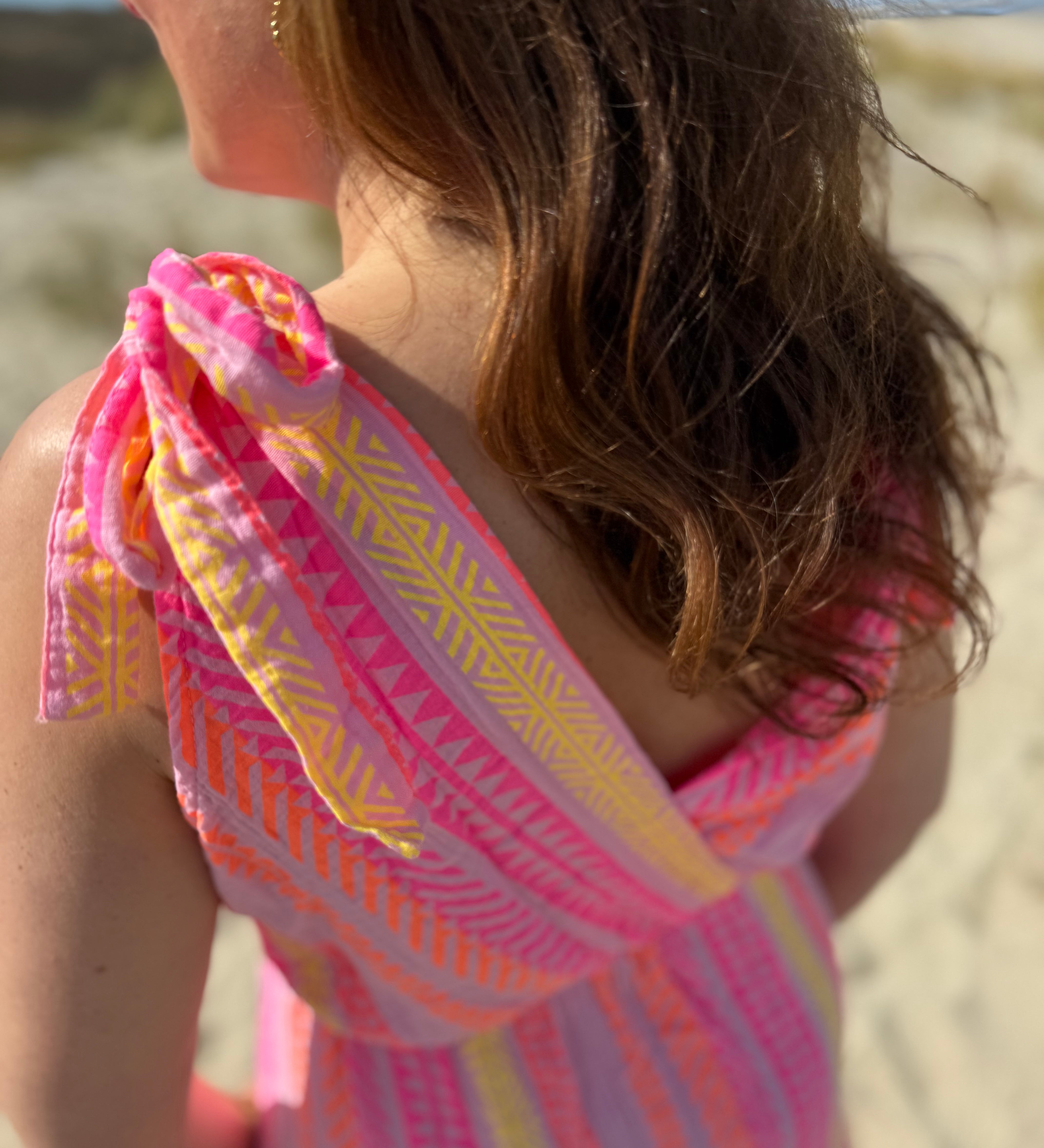 Musselinkleid in Midilänge mit V Ausschnitt. Es ist ärmellos und hat Schleifen an den Schultern zum binden. Das Kleid ist rosa mit pink, gelb und orangenen Ethno Muster.