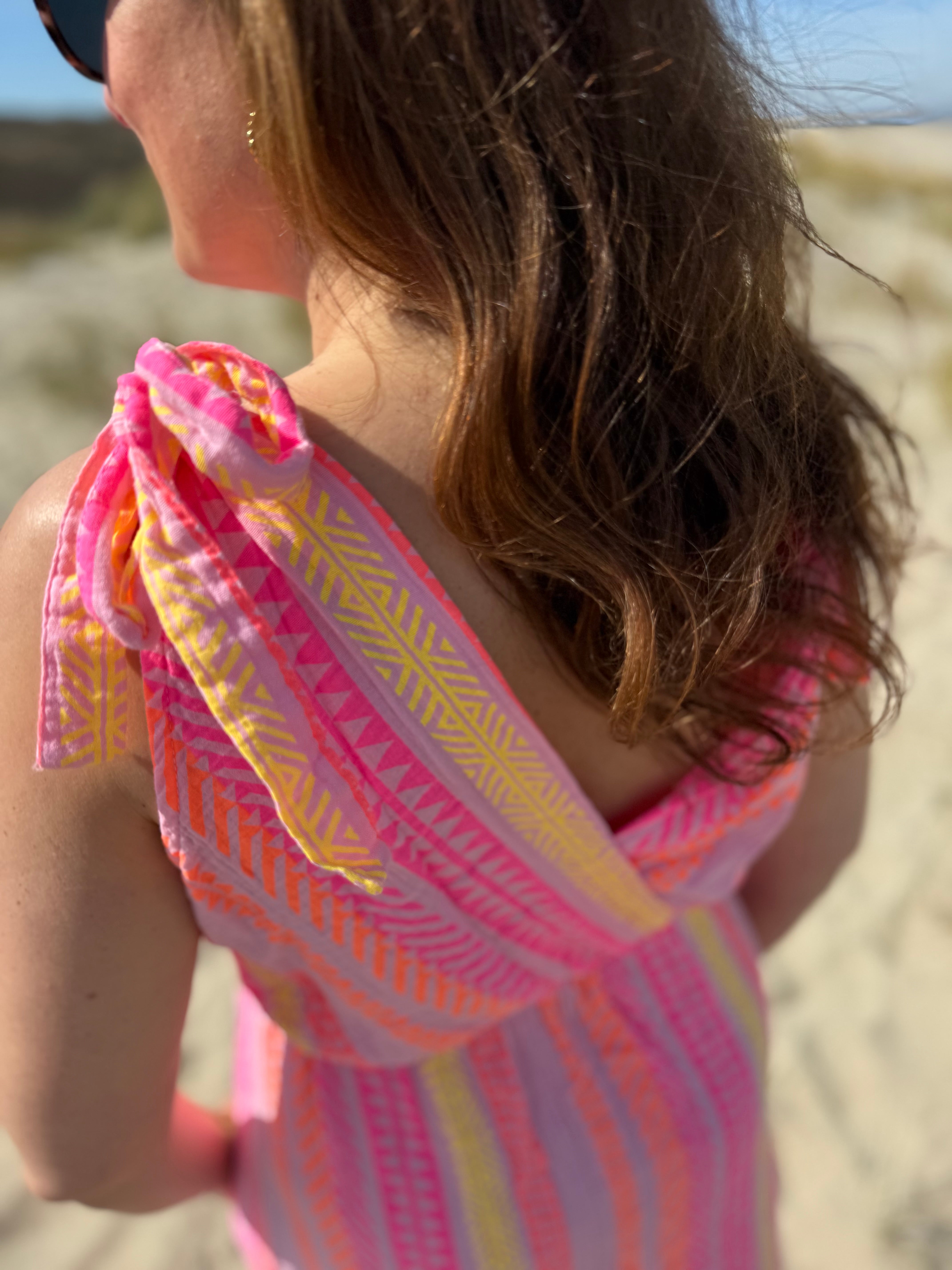 Musselinkleid in Midilänge mit V Ausschnitt. Es ist ärmellos und hat Schleifen an den Schultern zum binden. Das Kleid ist rosa mit pink, gelb und orangenen Ethno Muster.