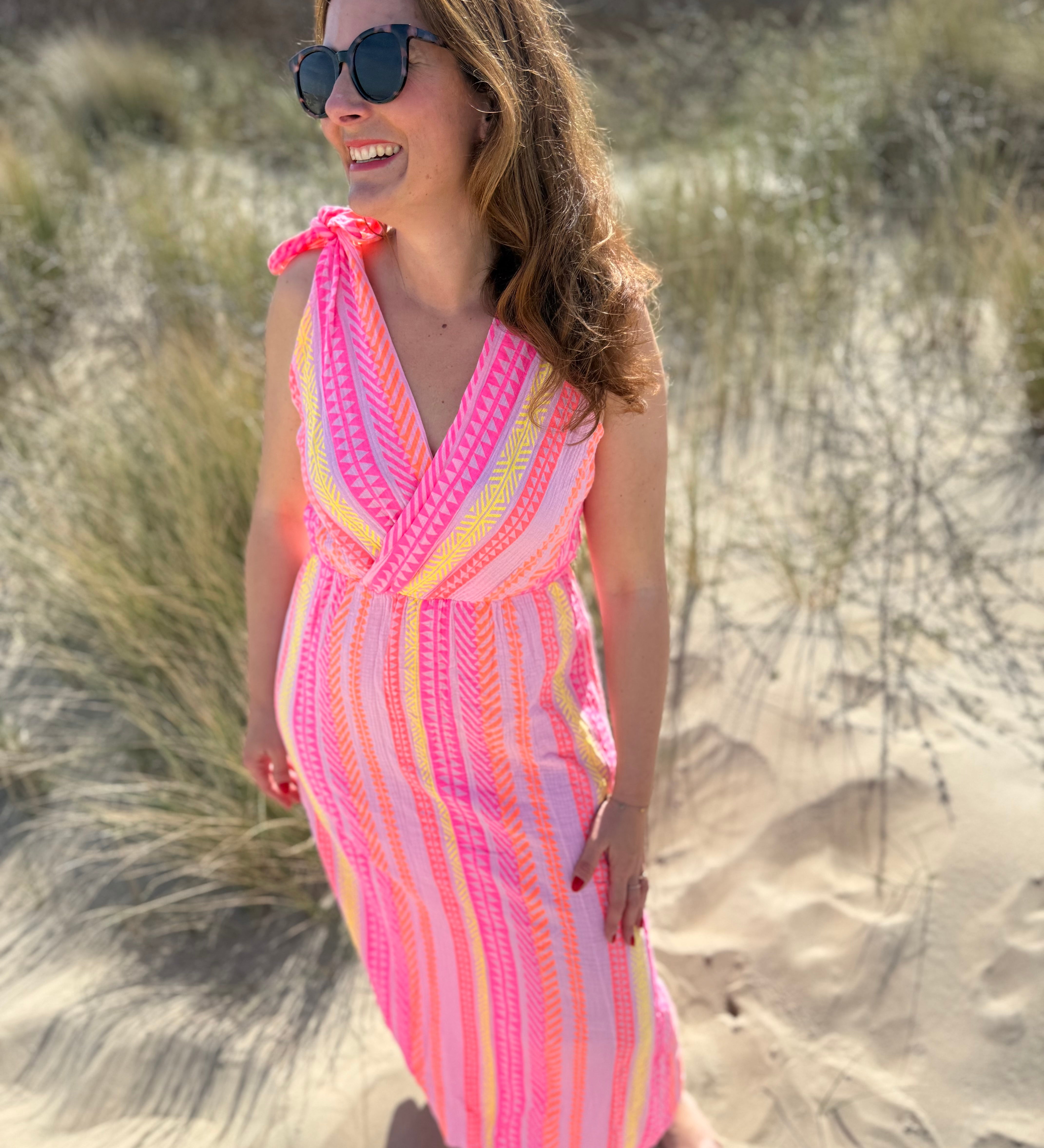Musselinkleid in Midilänge mit V Ausschnitt. Es ist ärmellos und hat Schleifen an den Schultern zum binden. Das Kleid ist rosa mit pink, gelb und orangenen Ethno Muster.