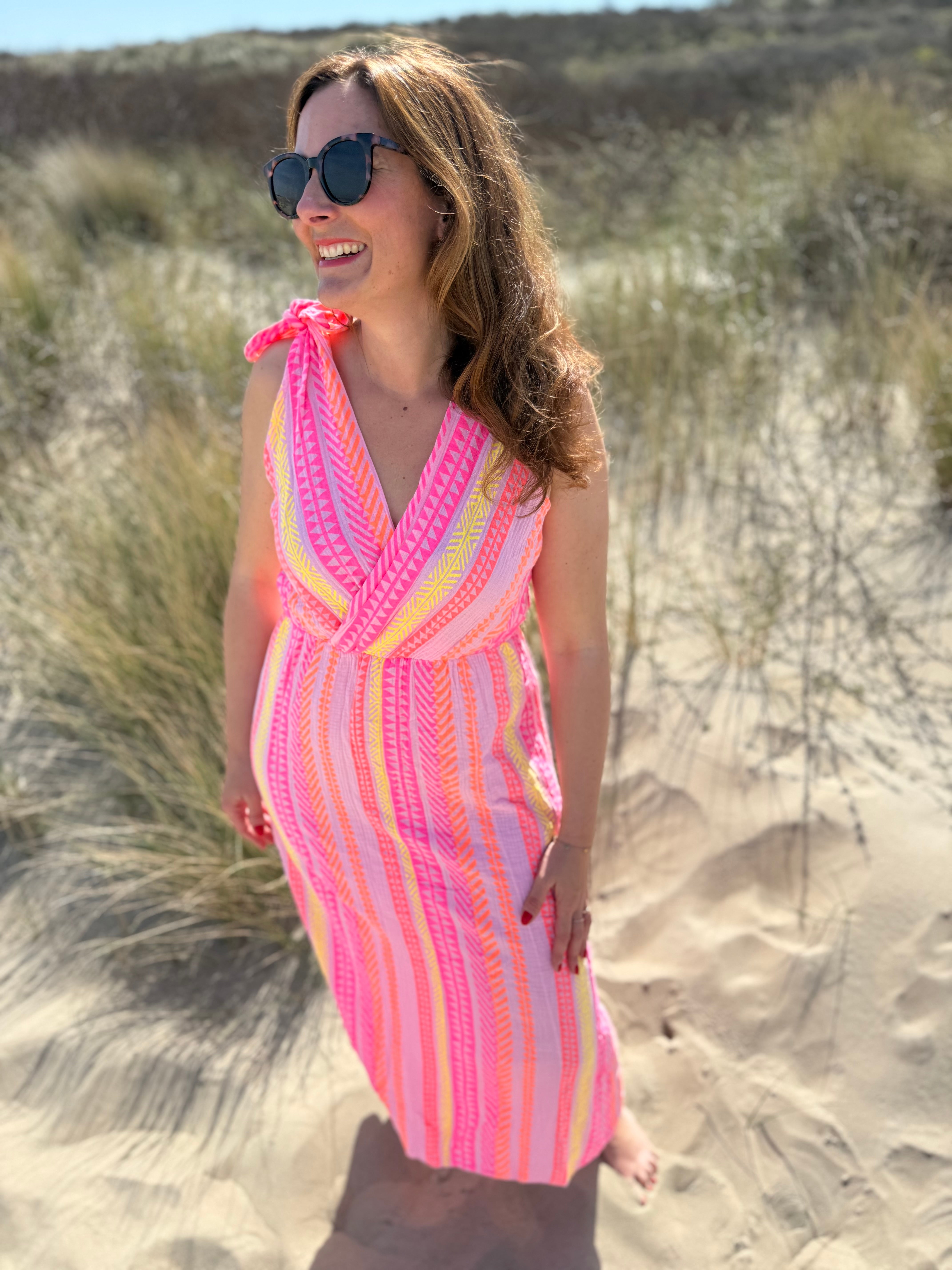Musselinkleid in Midilänge mit V Ausschnitt. Es ist ärmellos und hat Schleifen an den Schultern zum binden. Das Kleid ist rosa mit pink, gelb und orangenen Ethno Muster.