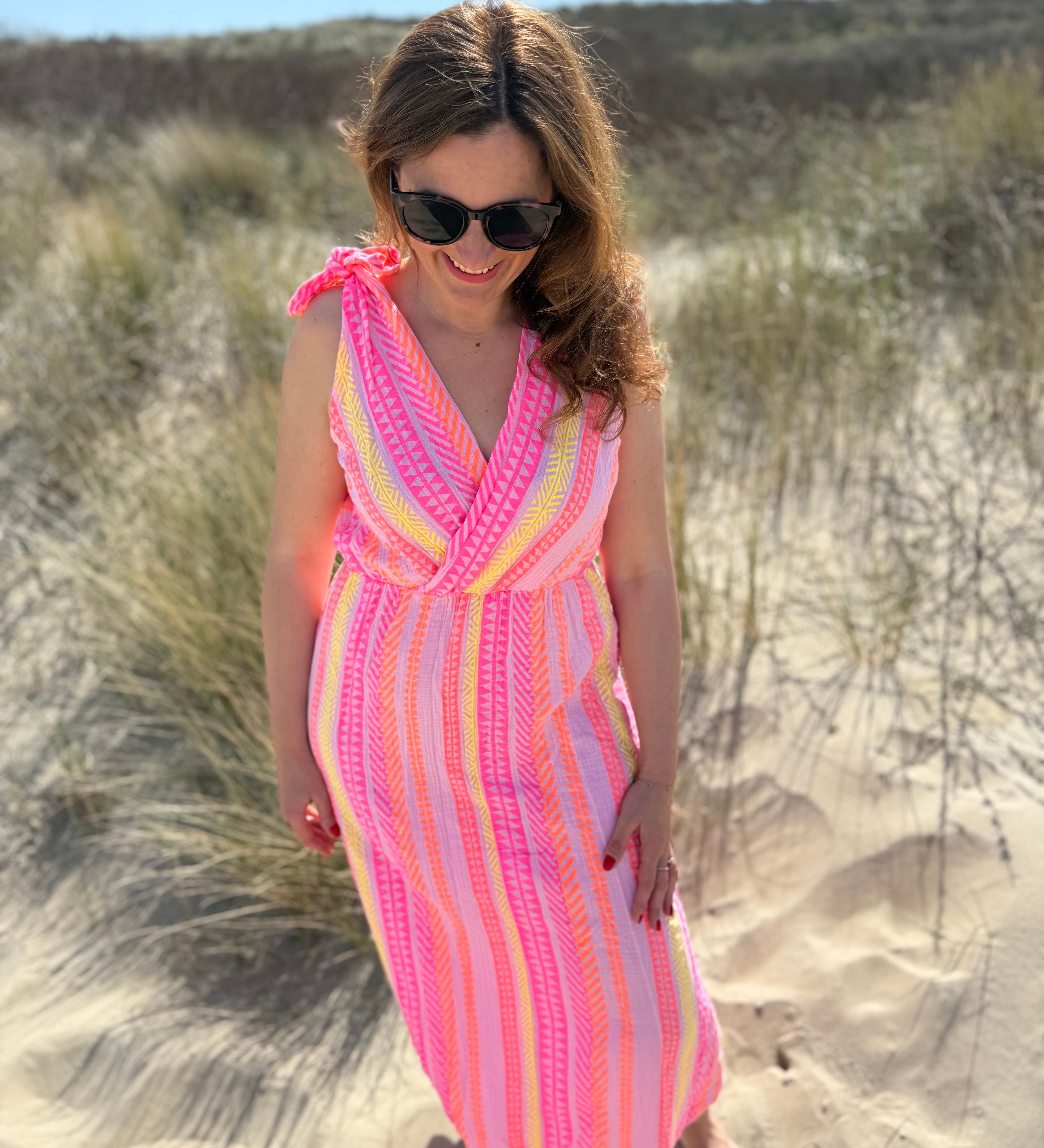 Musselinkleid in Midilänge mit V Ausschnitt. Es ist ärmellos und hat Schleifen an den Schultern zum binden. Das Kleid ist rosa mit pink, gelb und orangenen Ethno Muster.
